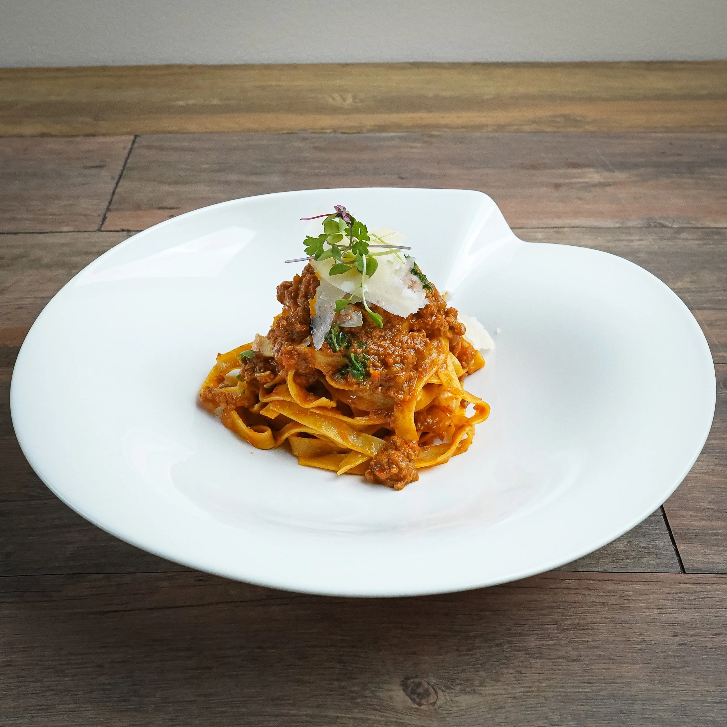 A plate of pasta with meat sauce garnished with microgreens and shaved cheese on a white dish.