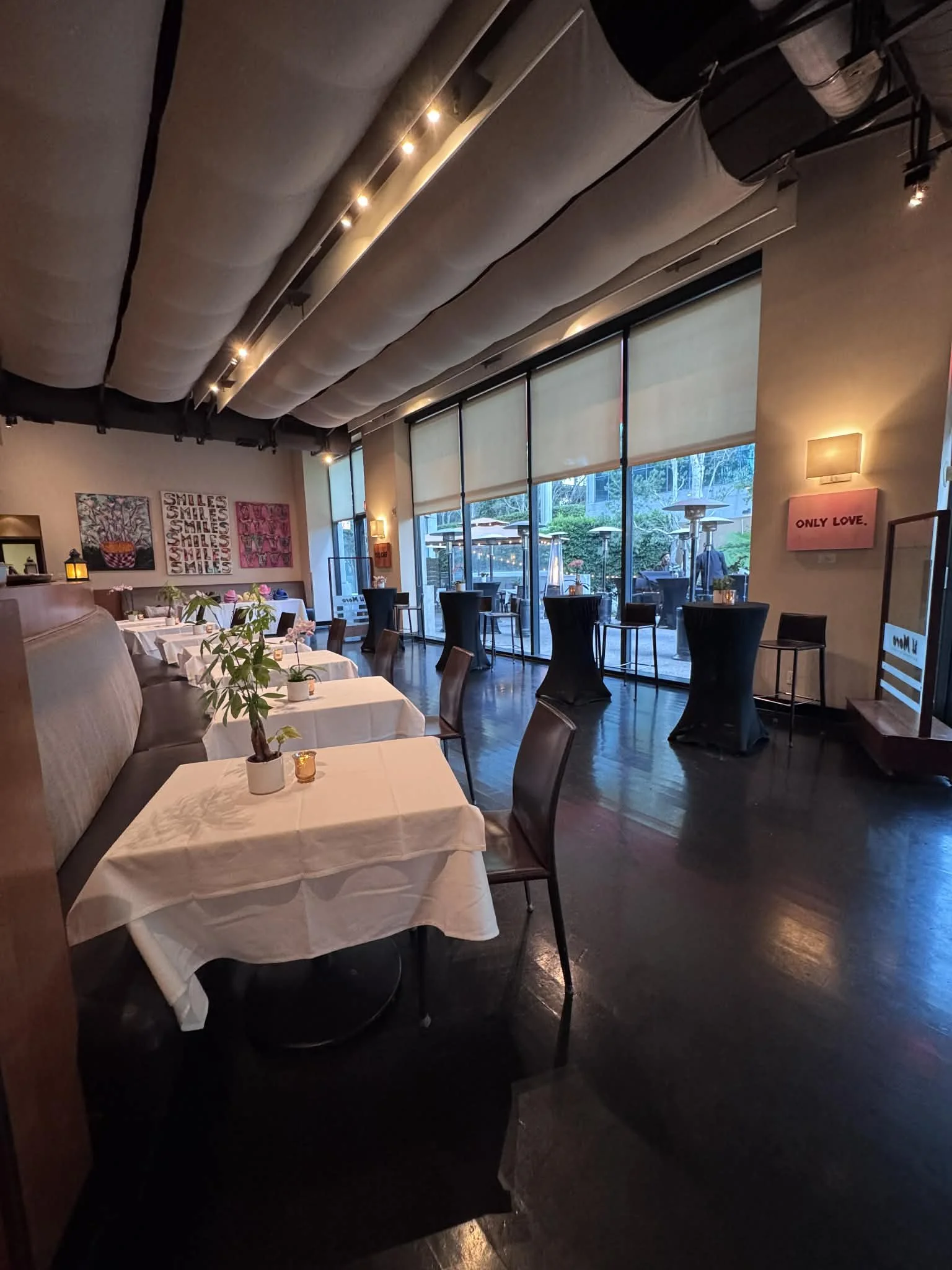 Inside a restaurant with white tablecloths, potted plants, and framed artwork on the walls. Large windows reveal an outdoor patio with tables, chairs, and umbrellas. Dim lighting creates a cozy atmosphere.