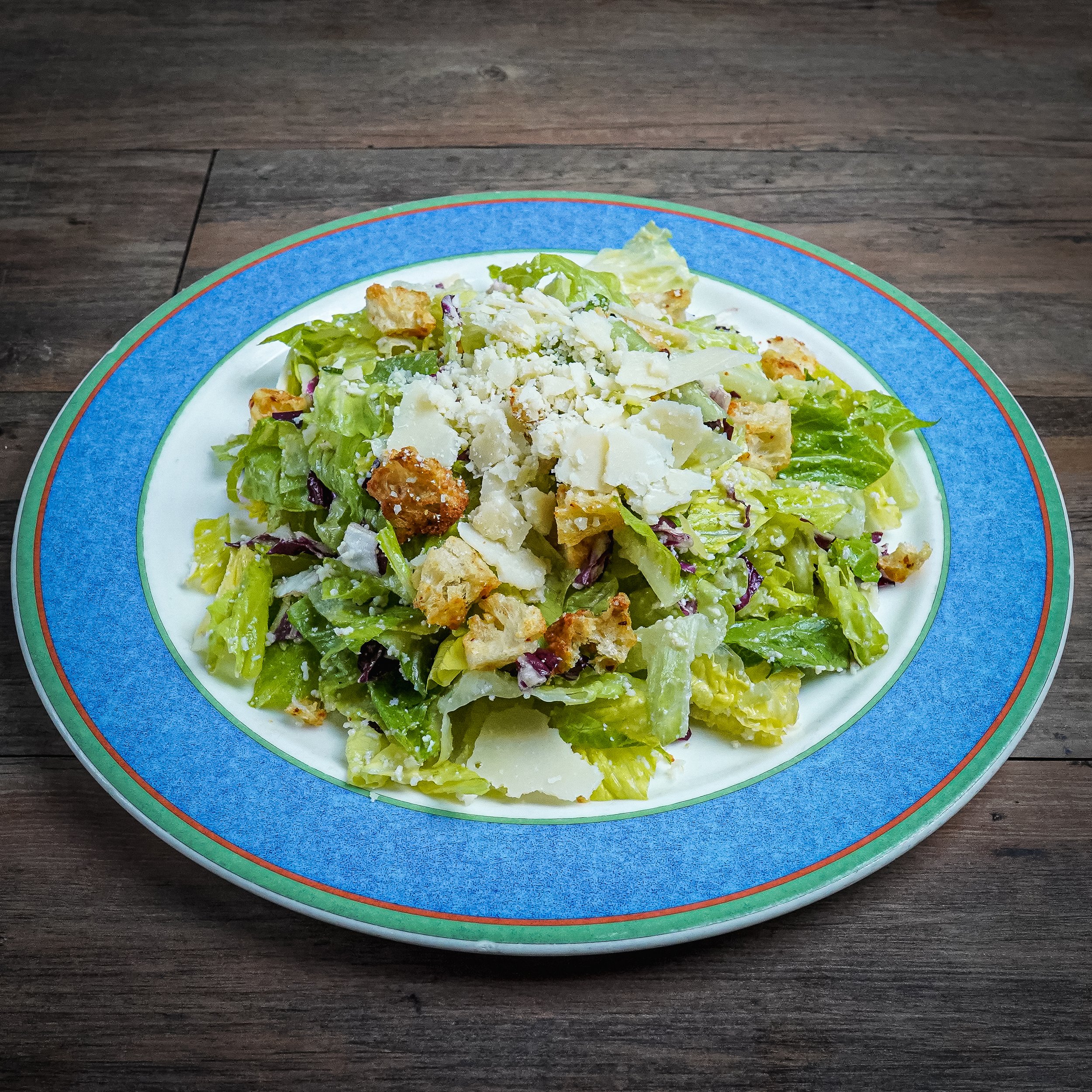 A Caesar salad on a decorative plate with chopped lettuce, croutons, grated cheese, and Caesar dressing, placed on a wooden table.