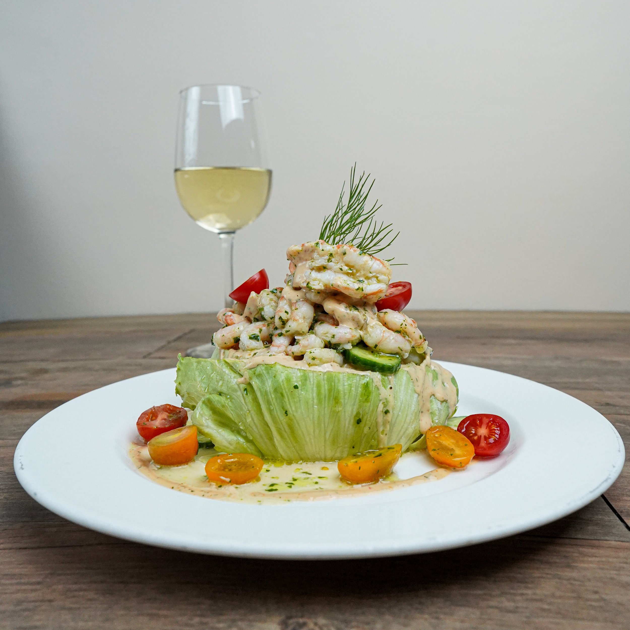 A plate of shrimp and vegetable salad served in a lettuce cup, garnished with cherry tomatoes and dill, with a glass of white wine in the background.