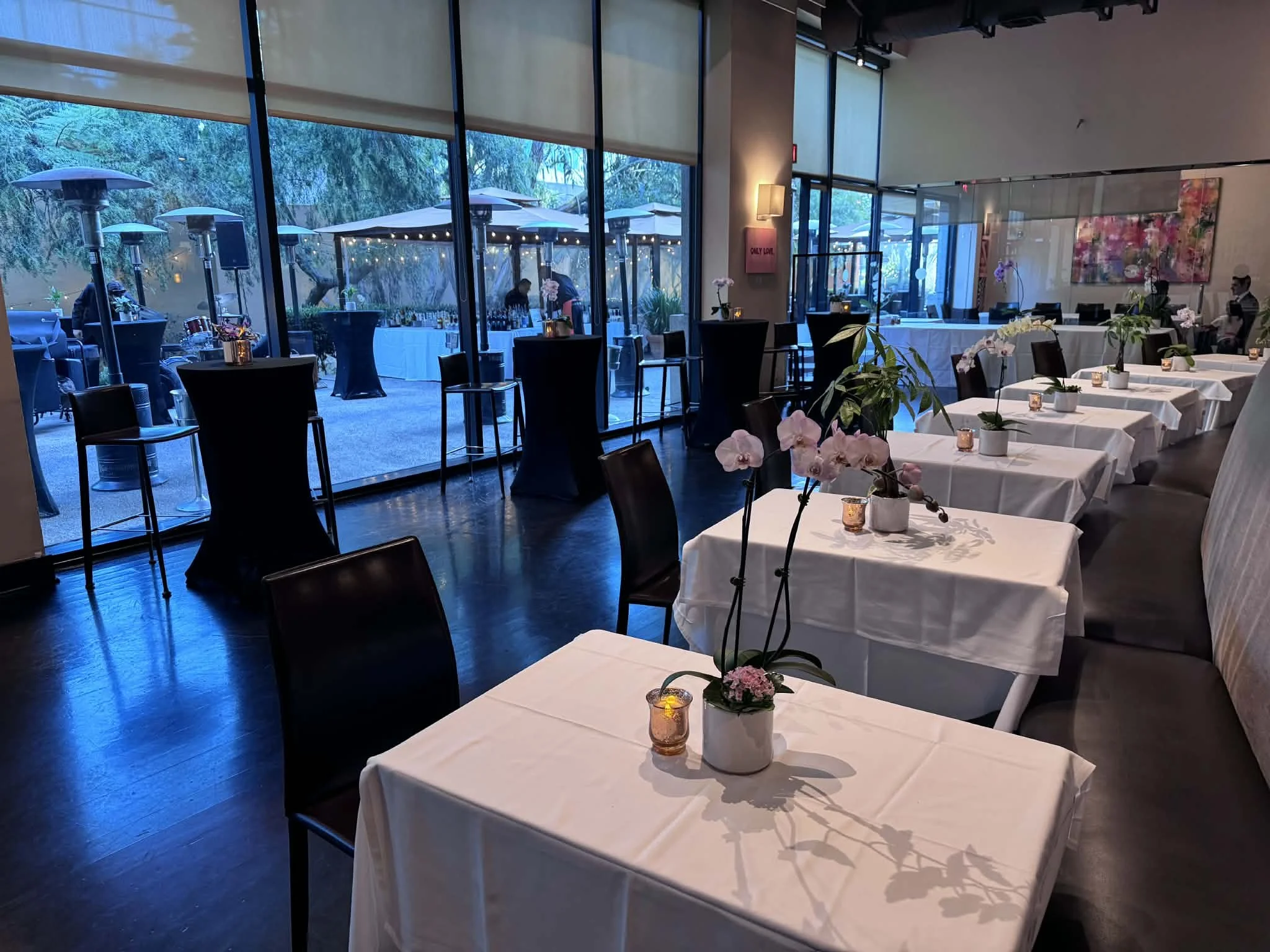 A dining area with tables covered in white tablecloths, decorated with potted orchids and candle holders. Large windows reveal an outdoor patio with umbrellas and string lights, and there are a few people inside and outside.