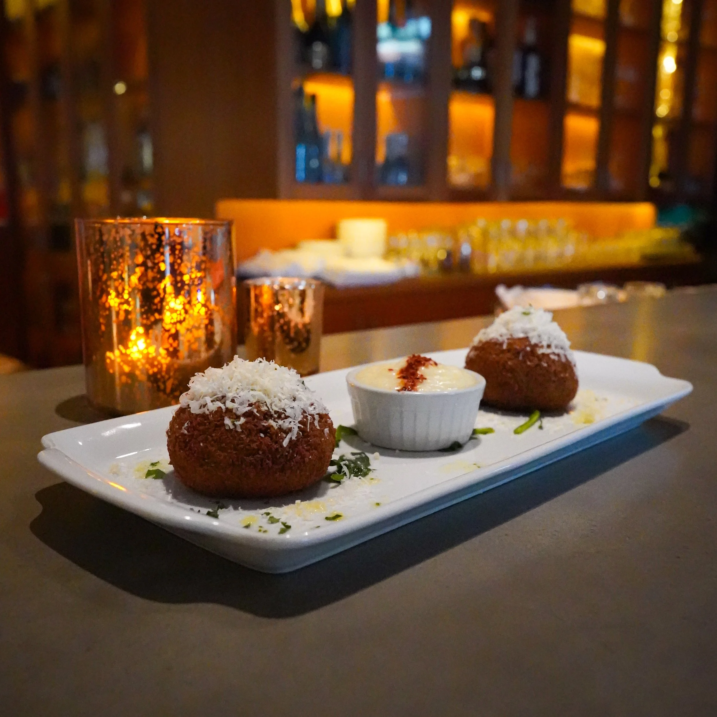 Plate with two croquettes topped with shredded cheese and a small ramekin of creamy dip in a dimly lit restaurant setting with candles in the background.