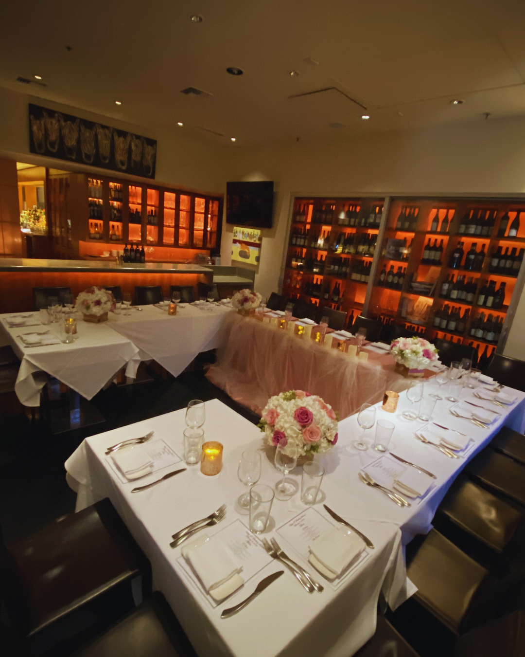 A restaurant or event space decorated for a wedding or celebration, with a U-shaped table arrangement, white tablecloths, floral centerpieces, candles, and place settings, against a backdrop of wine bottles on shelves, with warm lighting.