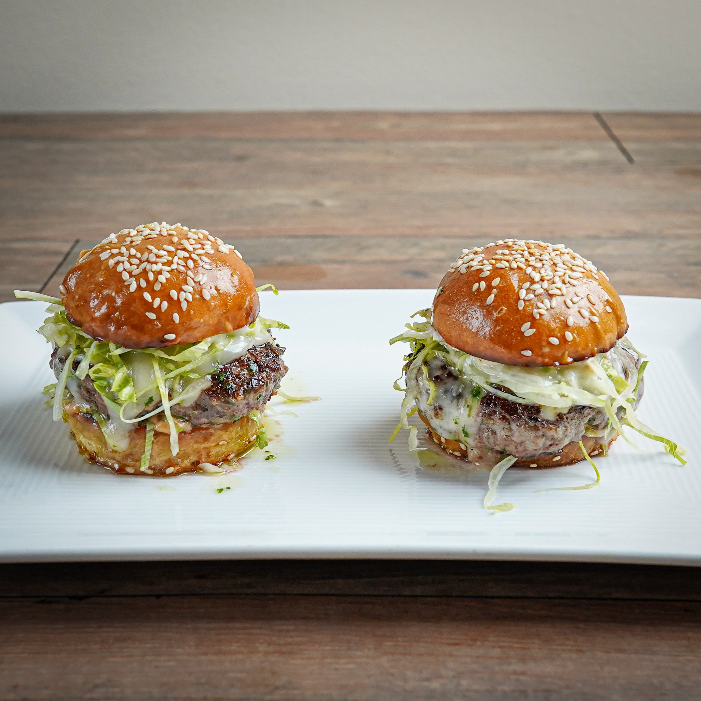 Two mini cheeseburgers with sesame seed buns, lettuce, and beef patties on a white rectangular plate.