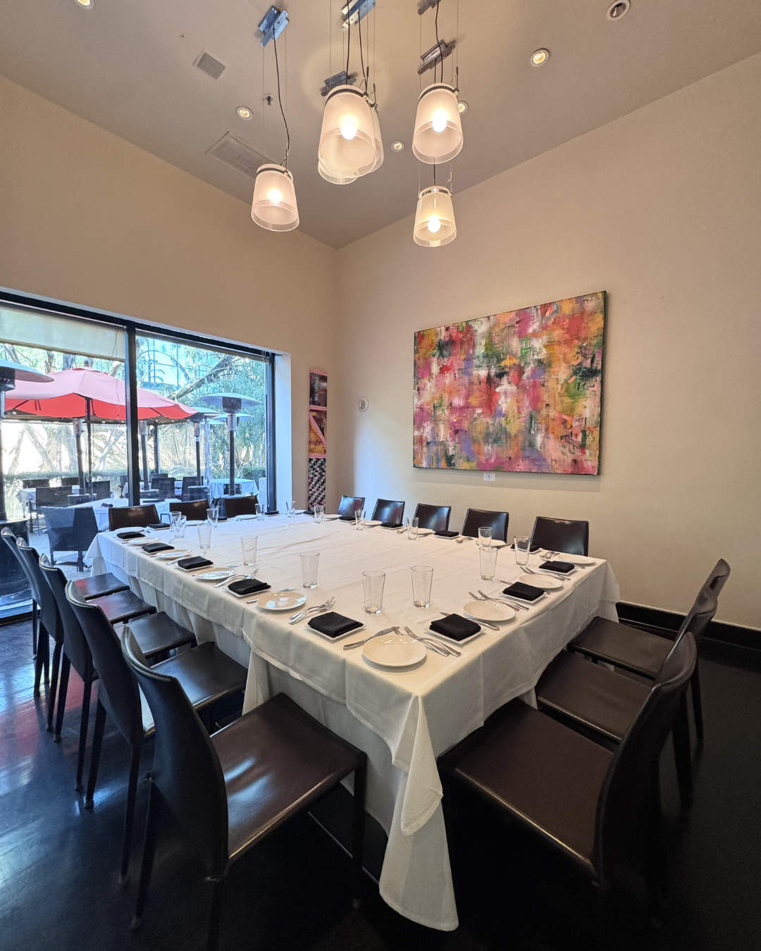 A dining room with a large rectangular table set for a meal, surrounded by black chairs. The table has white tablecloths, plates, glasses, and napkins. There is a colorful abstract painting on the wall and a large window with a view of an outdoor patio with red umbrellas and trees. Hanging pendant lights are above the table.