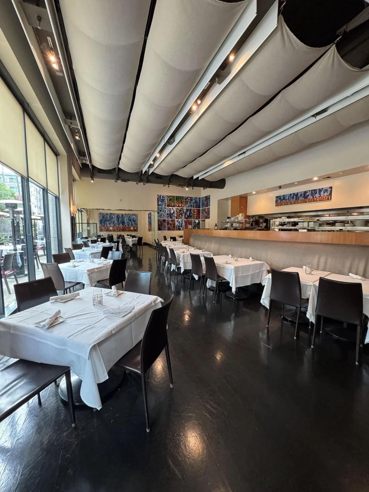 Interior of a modern restaurant with white tablecloths, black chairs, and colorful artwork on the walls, featuring large windows and a decorative ceiling.