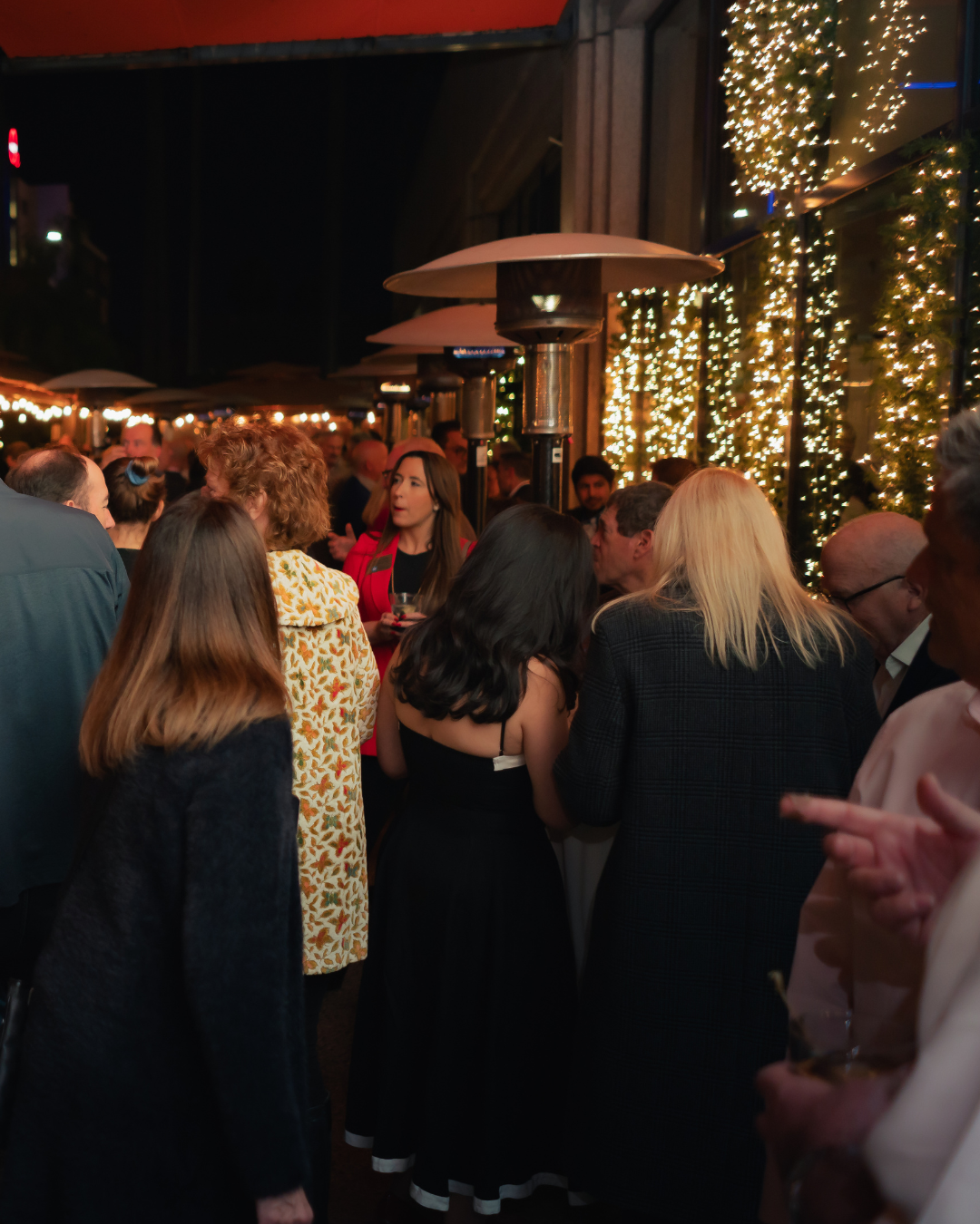 People gathered at an outdoor evening event with string lights and outdoor heaters.
