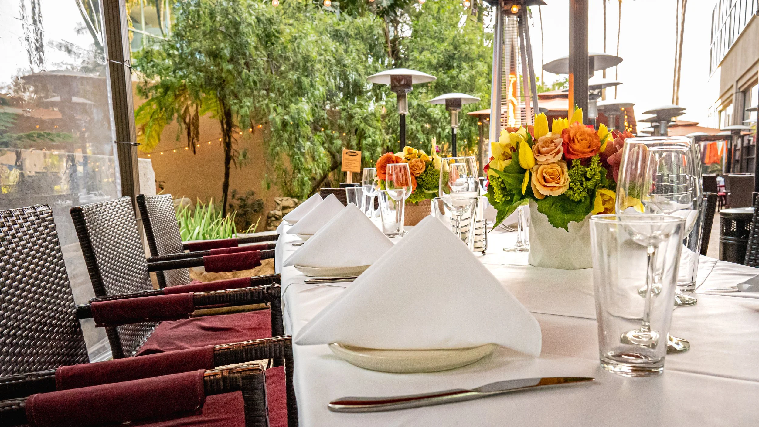A banquet table set with white tablecloths, folded napkins, wine glasses, and floral centerpieces. An outdoor patio with trees, string lights, and patio heaters in the background.