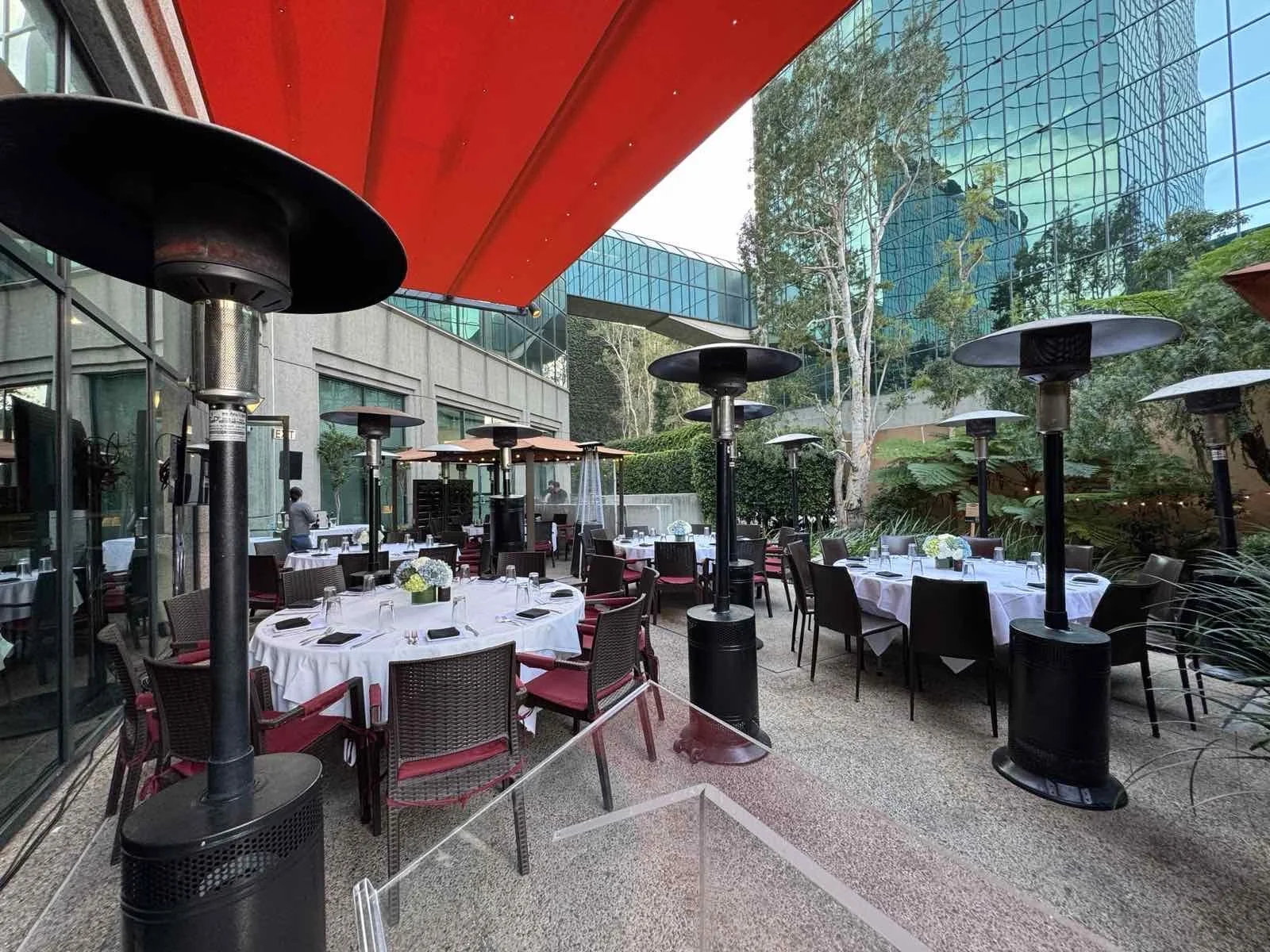 Outdoor restaurant patio with tables covered in white tablecloths, red cushioned chairs, and outdoor heaters, surrounded by greenery and modern glass buildings.