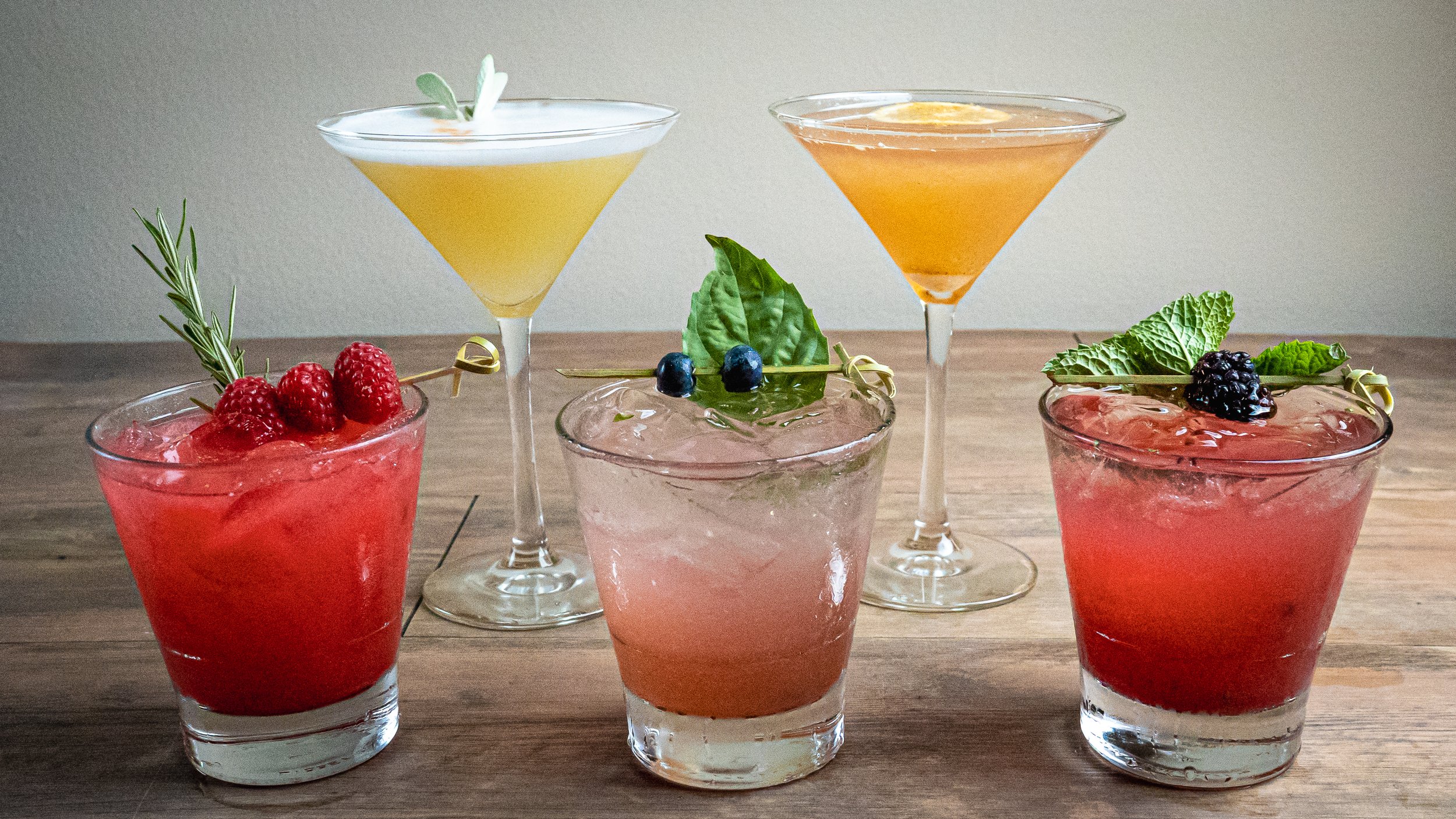 Six cocktails on a wooden surface, three in short glasses garnished with berries and herbs, and two in martini glasses with citrus slices.