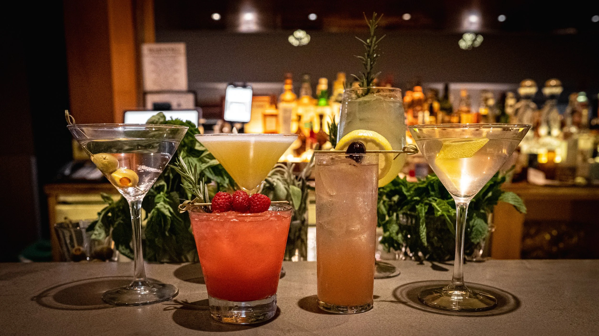 Five cocktails are displayed on a bar counter with a background of liquor bottles. The drinks include a martini, a yellow cocktail garnished with a lemon slice, a pink cocktail with raspberries, a tall cocktail with lemon slices and a cherry, and a lemon martini.