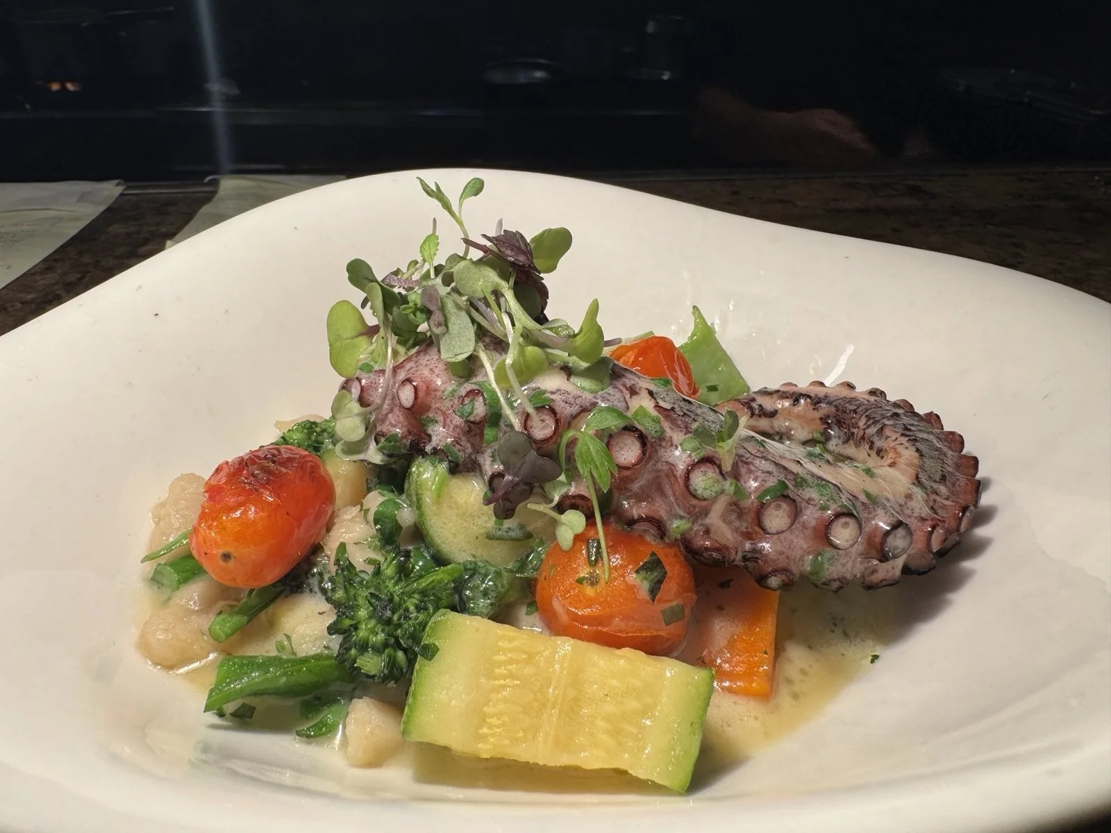 A plated dish featuring an octopus tentacle garnished with microgreens, alongside cherry tomatoes, sliced zucchini, broccoli, carrots, and a creamy sauce.