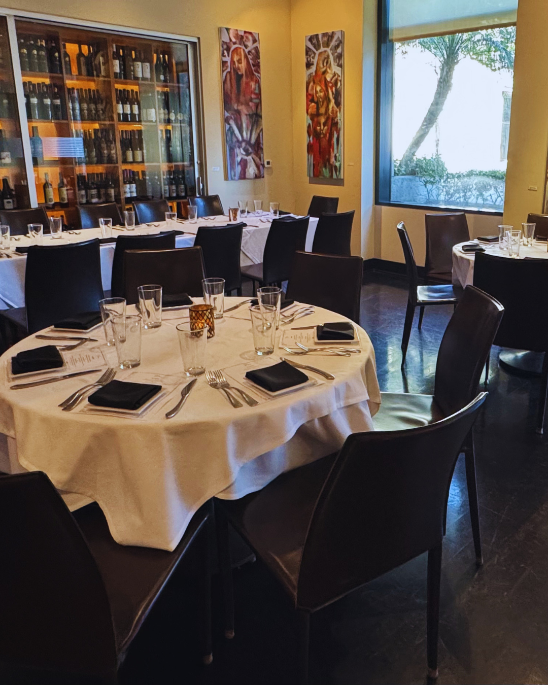 A restaurant dining room with round tables set with black napkins, menus, glasses, and silverware, and paintings on the wall near a large window showing outdoor greenery.