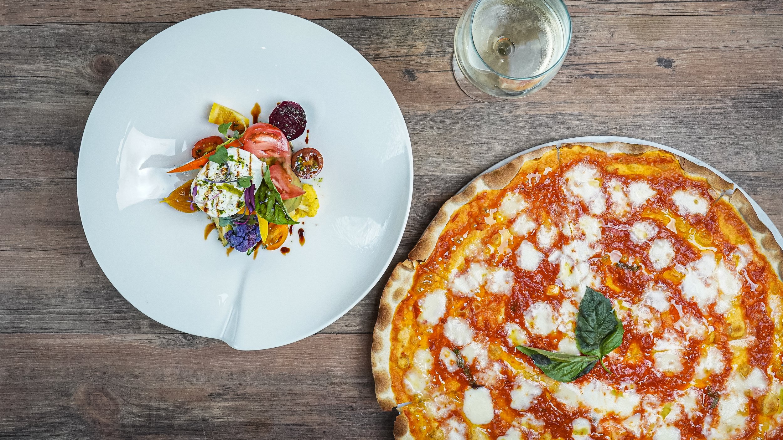 A top-down view of a wooden table with a slice of cheese pizza garnished with basil, a white plate with a colorful salad including various vegetables and herbs, and a glass of white wine.