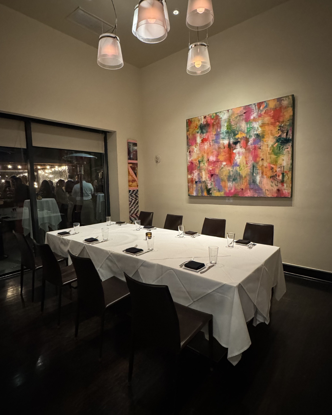 A restaurant dining room with a long rectangular table covered with a white tablecloth, set with glassware, napkins, and utensils, surrounded by dark chairs, with a colorful abstract painting on the wall, and a large window or sliding door showing people gathered indoors.
