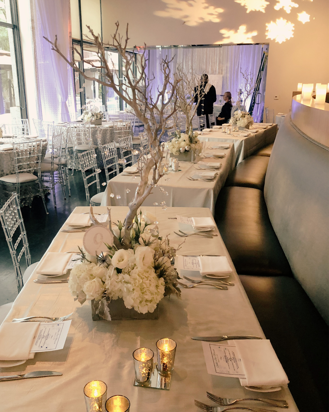 Elegant banquet table setup with white floral arrangements, candles, and place settings in a decorated event space with natural light.