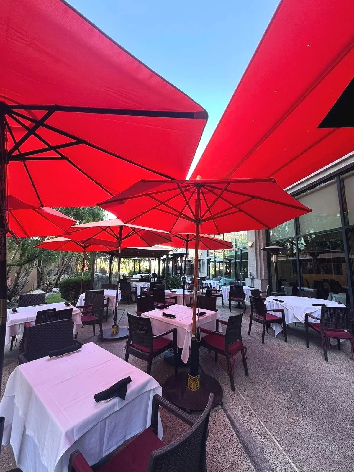 An outdoor restaurant patio with white tablecloths, black chairs with red cushions, and large red umbrellas providing shade.
