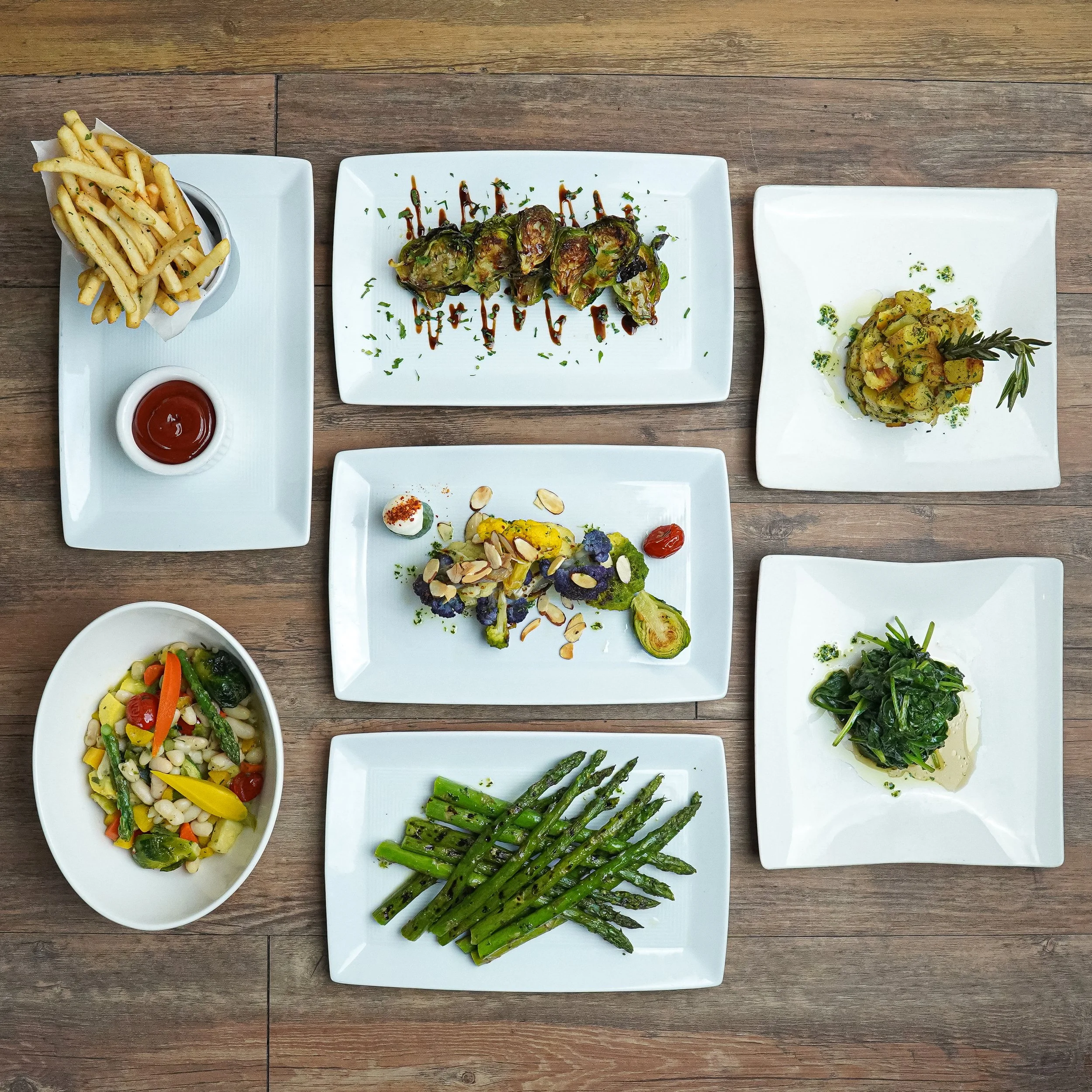 Assorted dishes of vegetables and appetizers on white plates and bowls, including french fries with ketchup, roasted Brussels sprouts, a mixed vegetable salad, grilled asparagus, and sautéed spinach, on a wooden table.