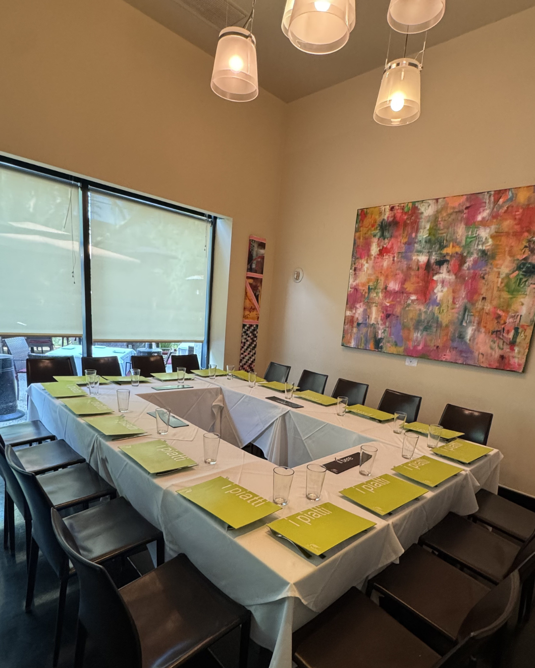 A conference room with a U-shaped table, set with glasses, black notebooks, and green pamphlets. Large window with partially closed beige blinds, colorful abstract painting on the wall, and modern ceiling lights.
