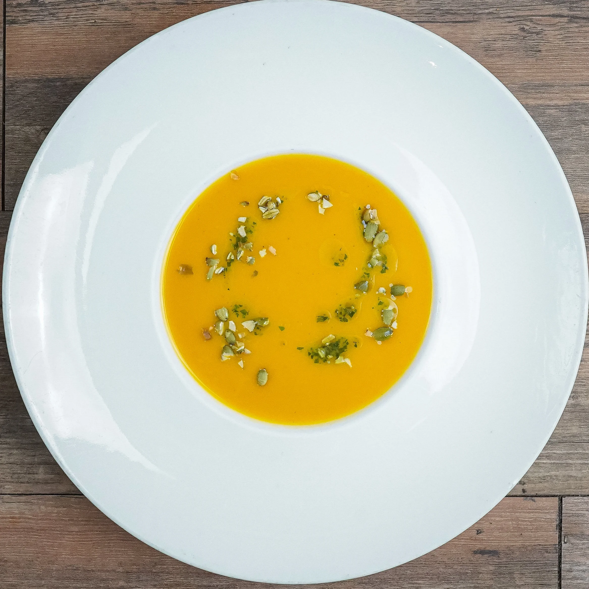 A bowl of creamy orange soup garnished with chopped herbs and pumpkin seeds on a white plate.