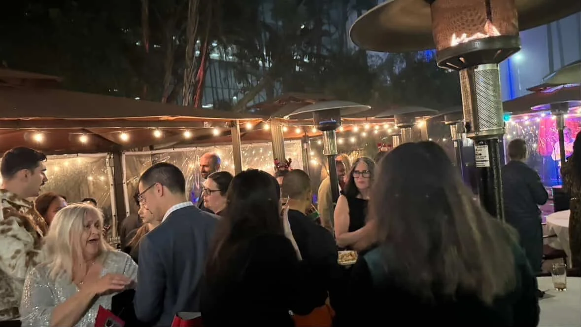 People socializing at an outdoor party or gathering under string lights and heaters with a decorated tent area.