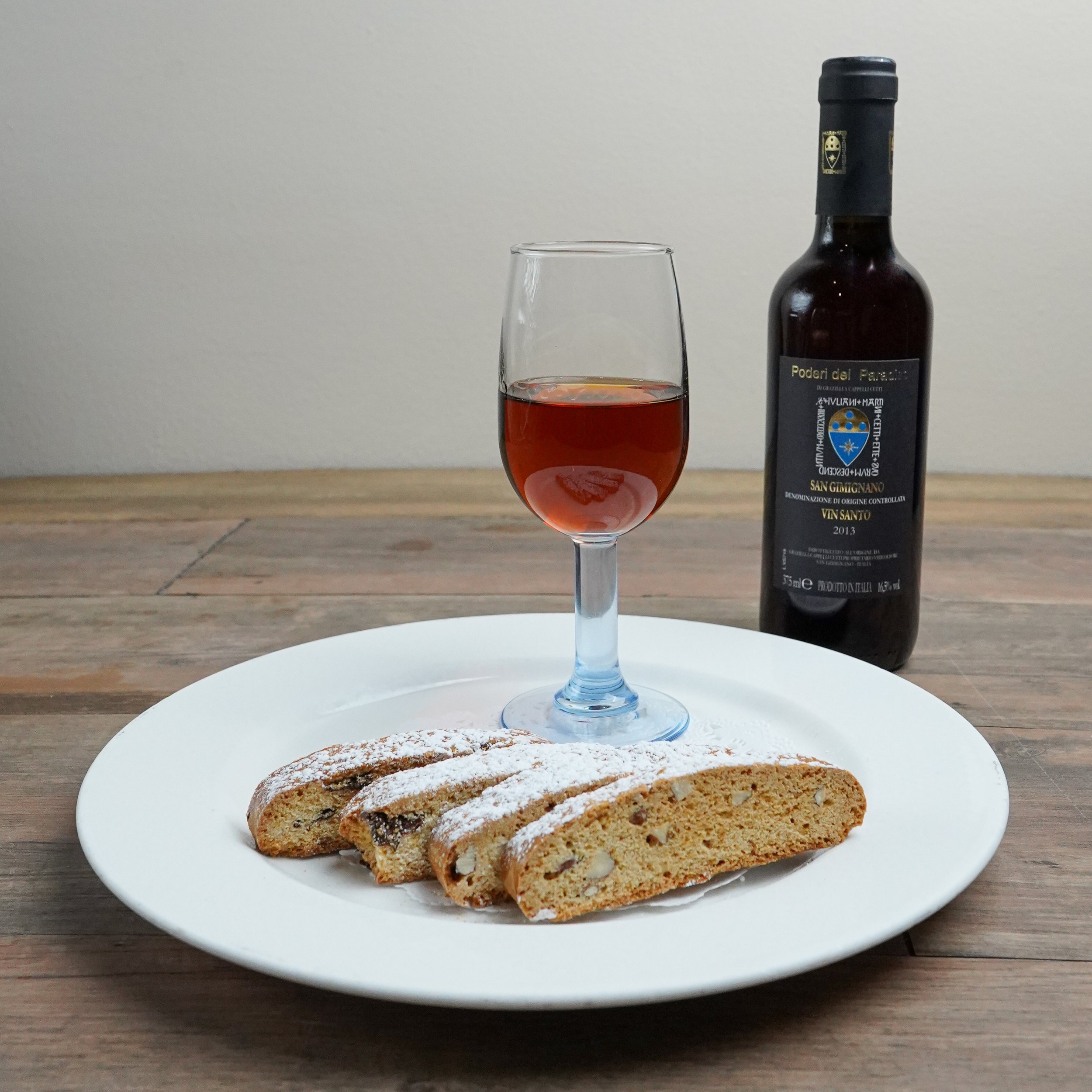 A glass of red wine on a white plate with biscotti cookies, and a bottle of red wine behind them on a wooden table.