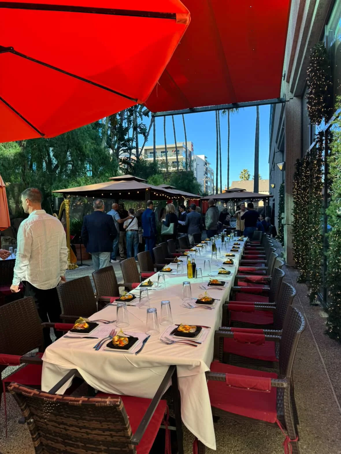 Long dining table set for a gathering with plates, glasses, and utensils, outside under umbrellas, with people mingling in the background and some trees and buildings nearby.