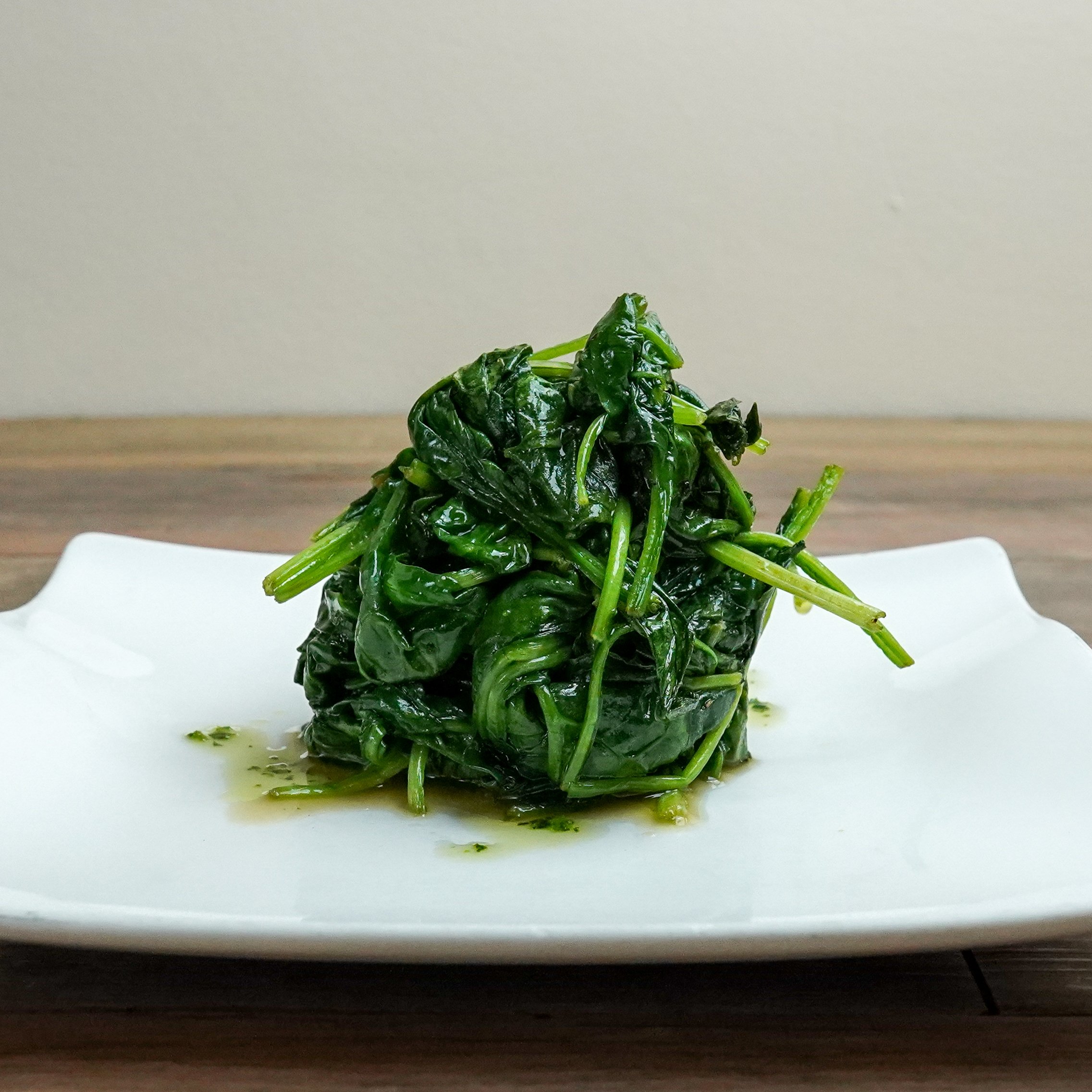 A white plate holding a serving of cooked spinach with a glossy appearance, garnished with some liquid.