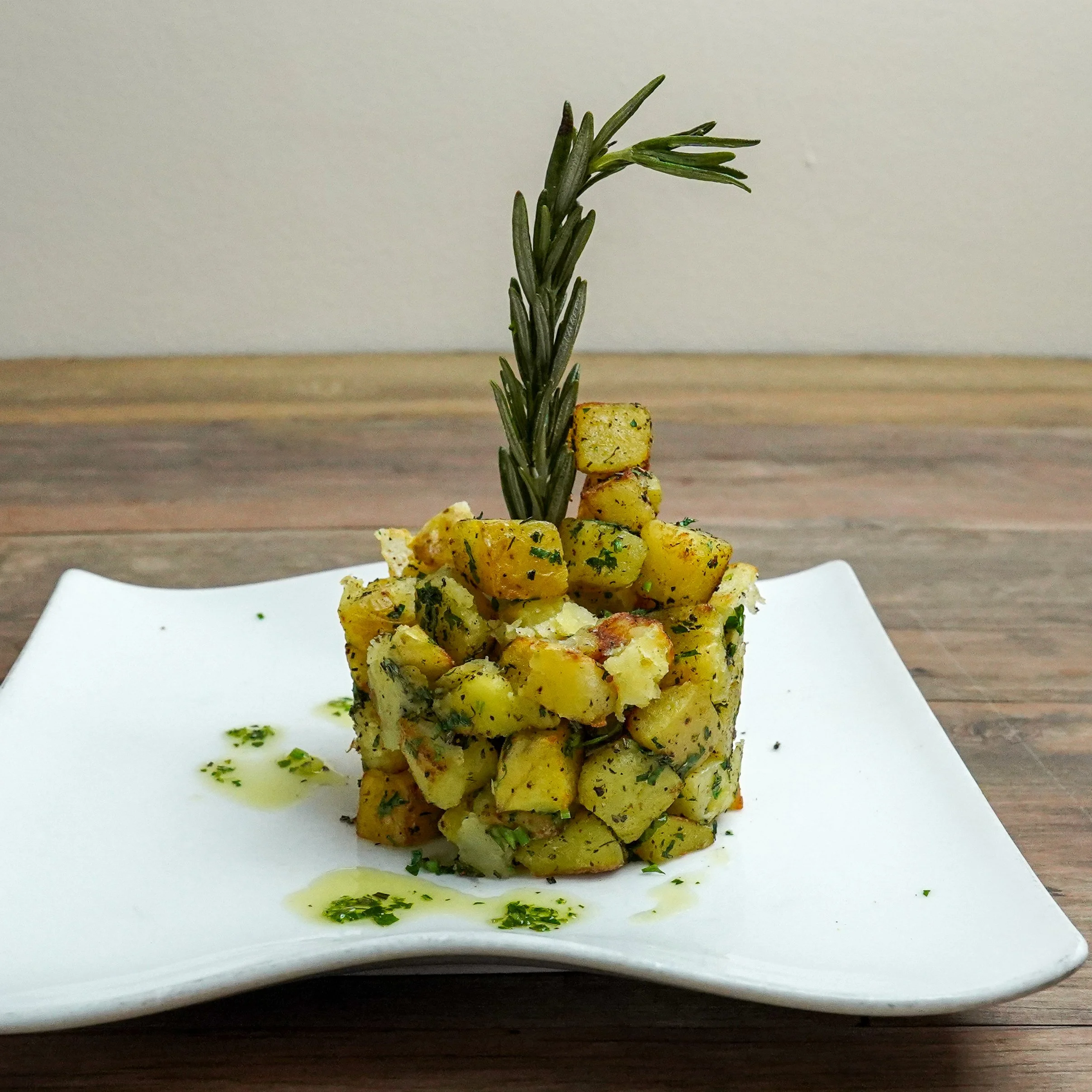 A white square plate holding a tower of a potato salad garnished with a sprig of rosemary, with some herbs and oil around the base.