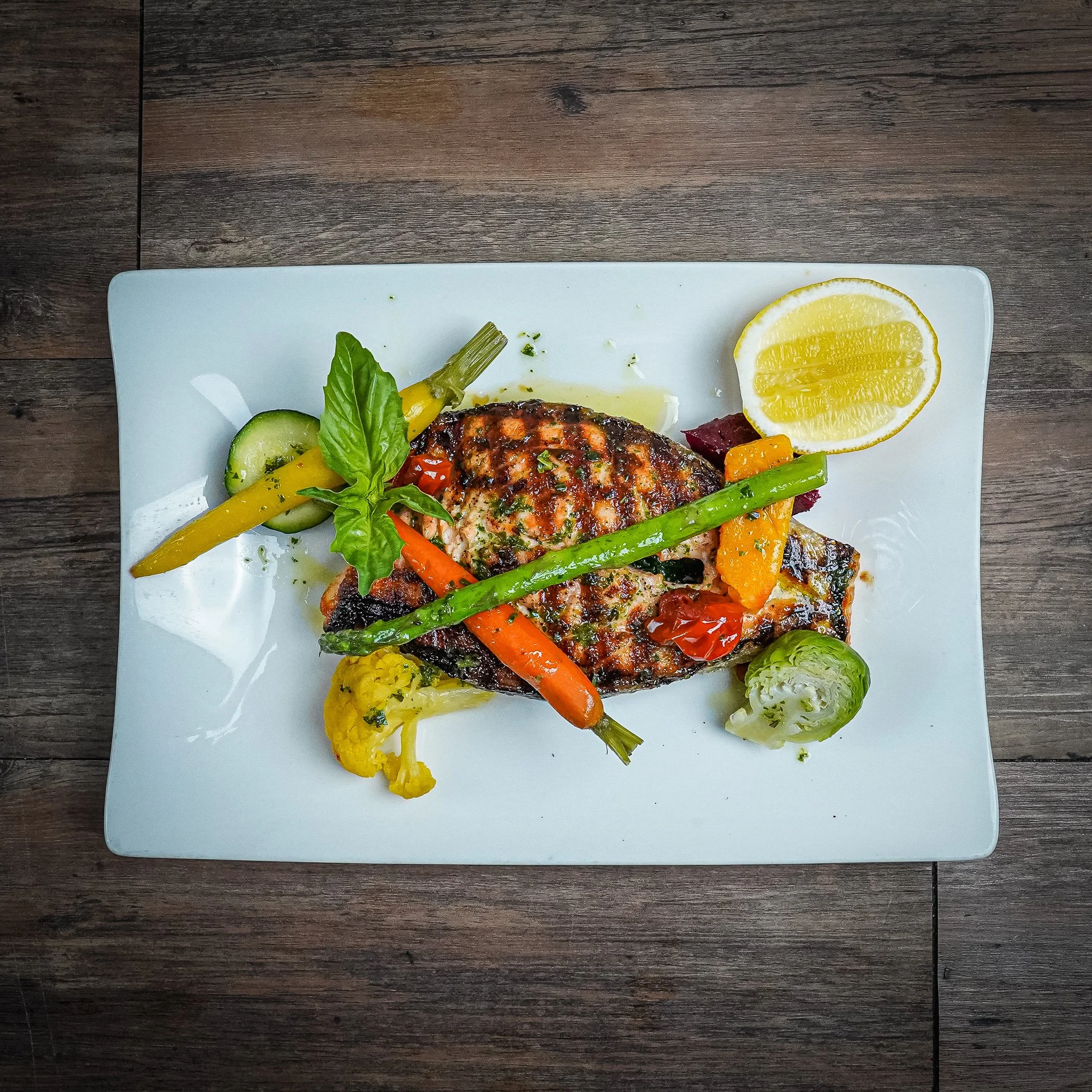 Grilled fish with assorted vegetables, lemon wedge, and garnish on a white rectangular plate.