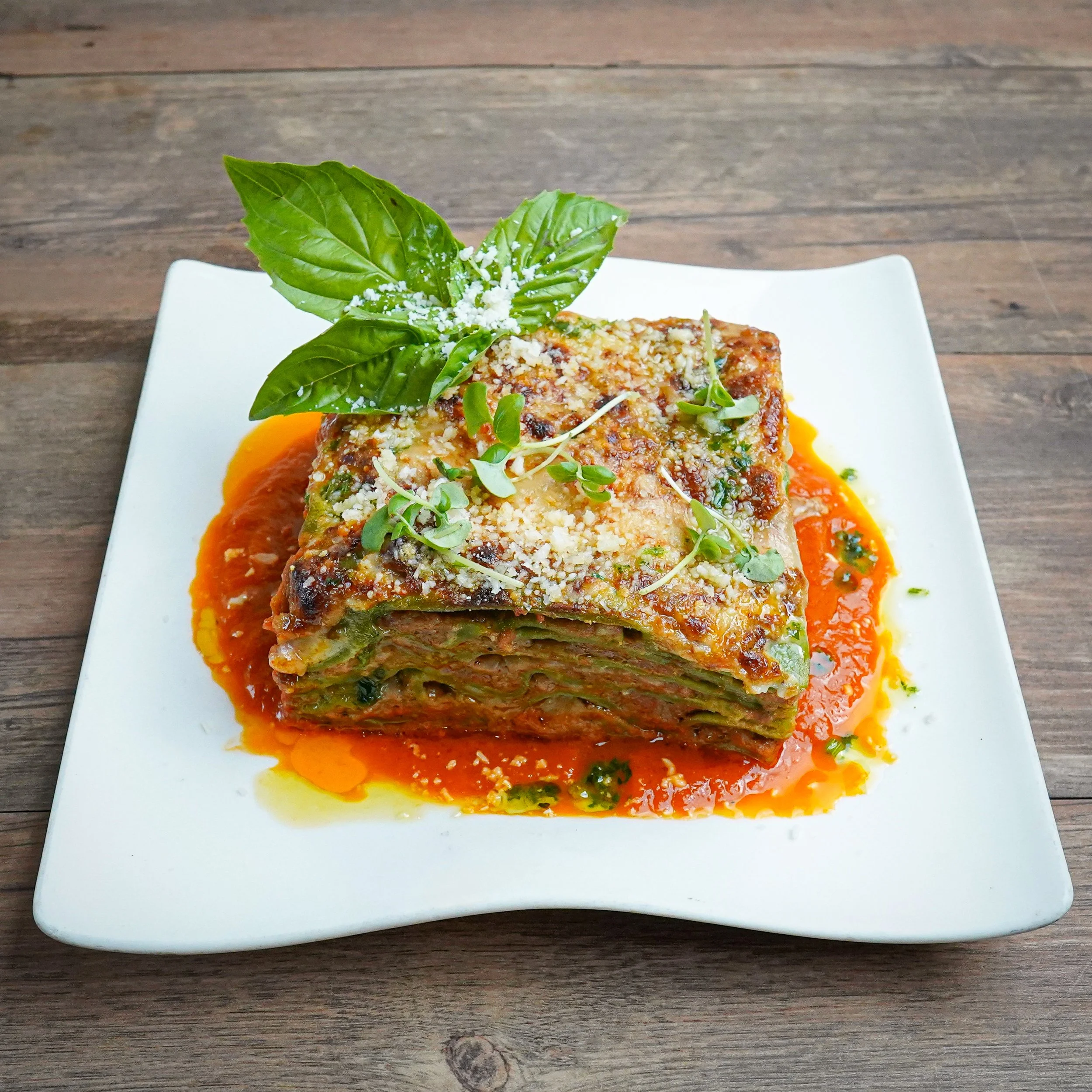 A plate of lasagna garnished with fresh basil leaves, microgreens, and grated cheese, served on a square white plate with tomato sauce.