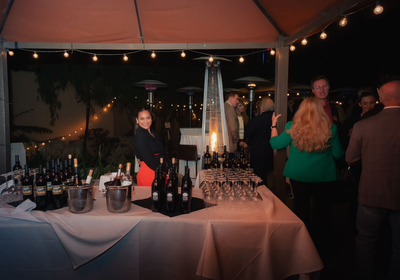 A woman smiling at a wine tasting event under a tent at night, with bottles of wine and empty glasses on the table, surrounded by guests.