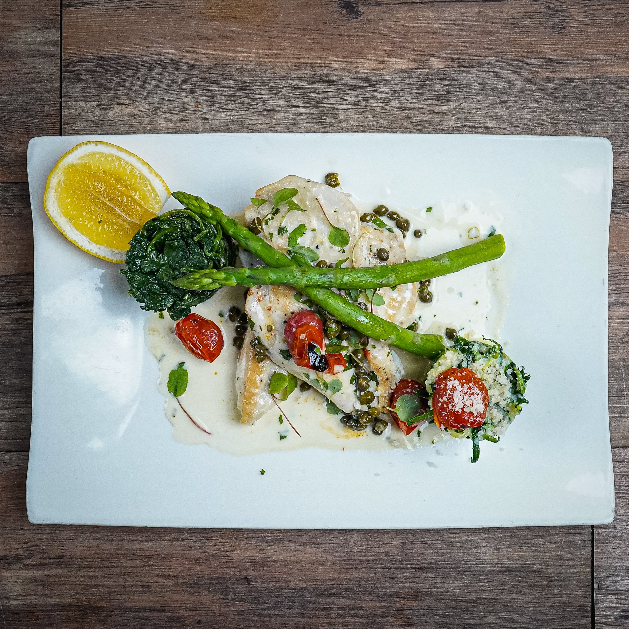 A plated dish with chicken, asparagus, cherry tomatoes, broccoli, lemon wedge, and a creamy white sauce, garnished with herbs and capers.