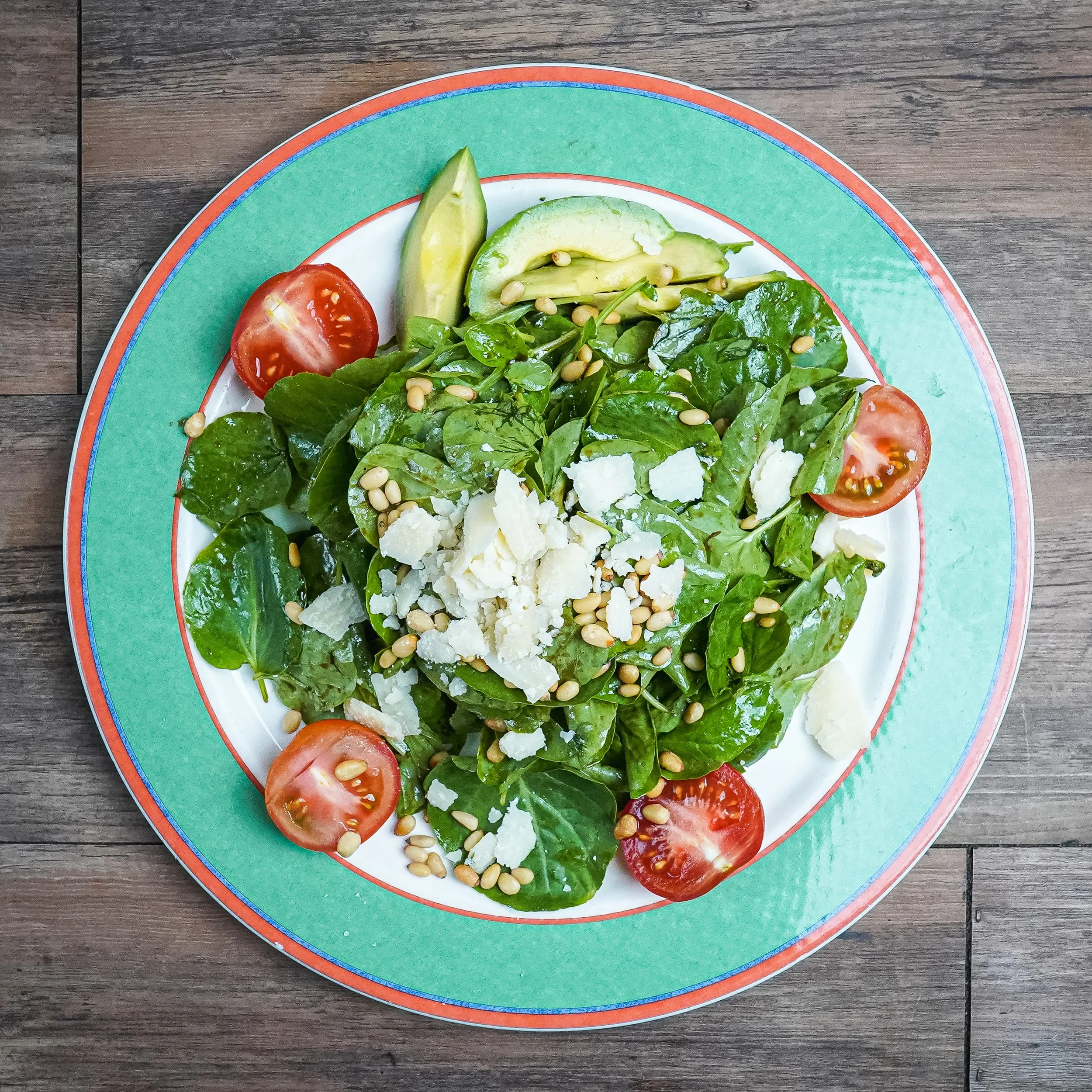 Mixed green salad with cherry tomatoes, avocado slices, shaved cheese, and pine nuts on a colorful plate.