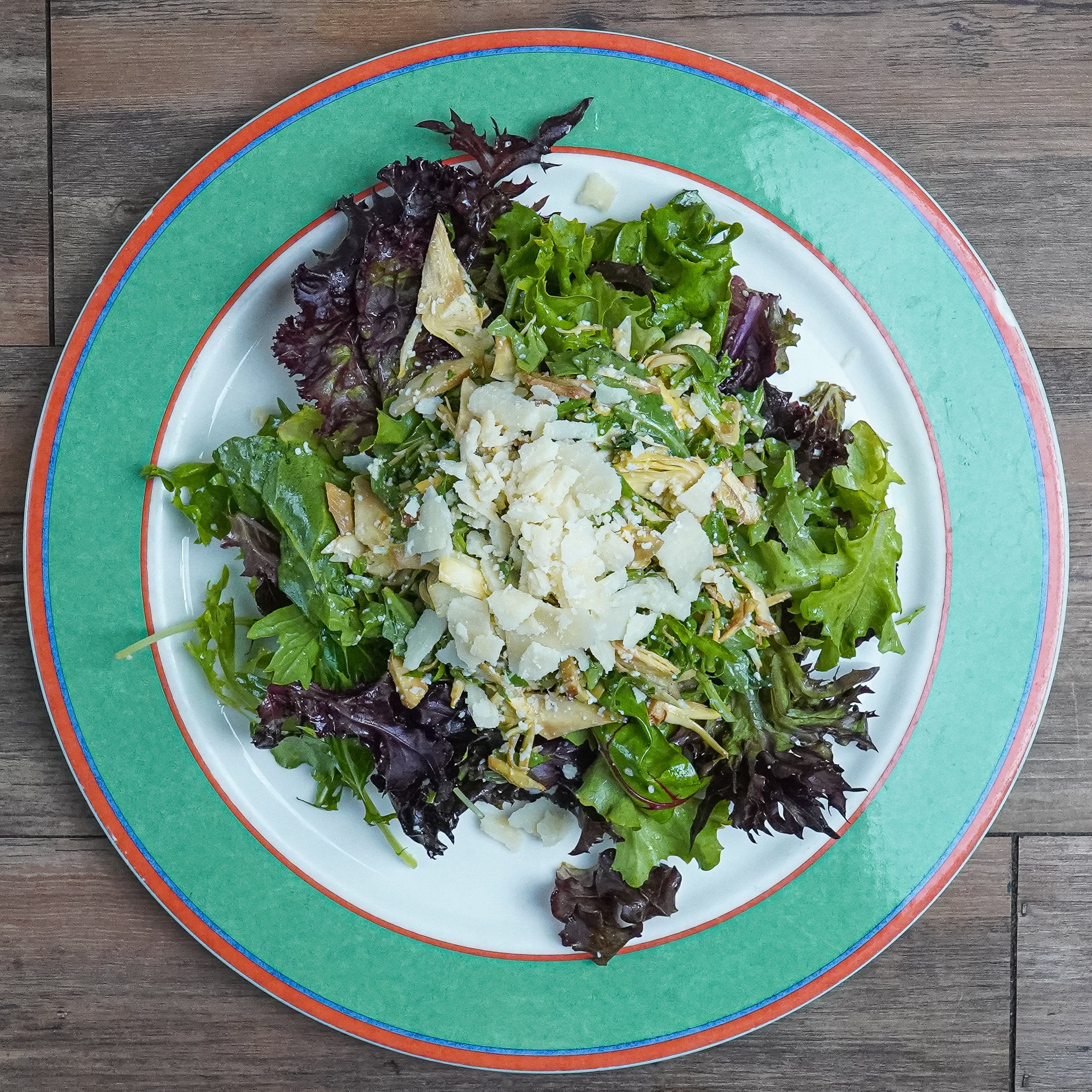 Mixed green salad topped with crumbled cheese on a white plate with a colorful green, red, and blue rim, placed on a wooden table.