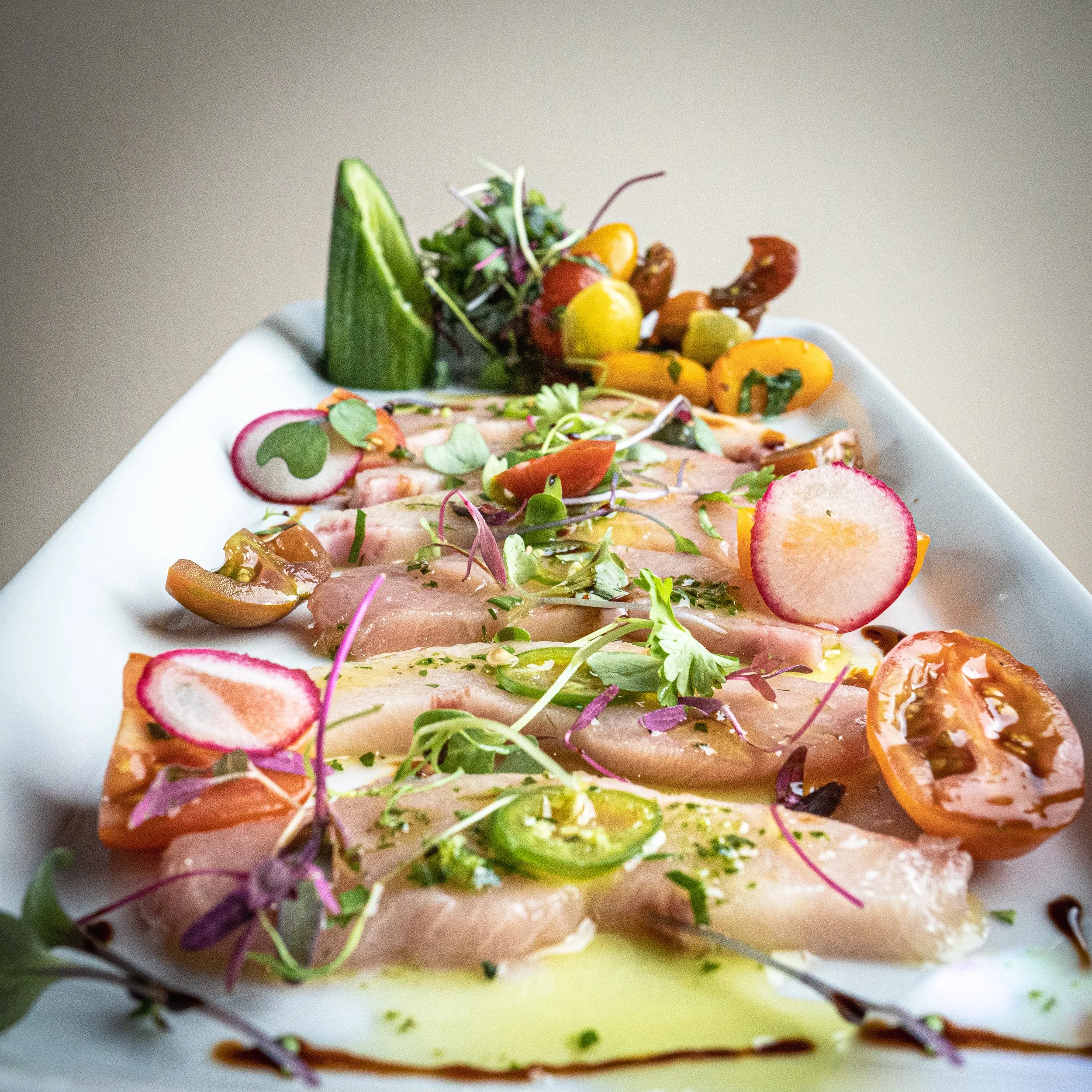 A white platter of raw fish carpaccio garnished with radish slices, microgreens, cherry tomatoes, sliced jalapeños, and a side of mixed vegetables including cherry tomatoes, yellow and green peppers, and cucumber slices.