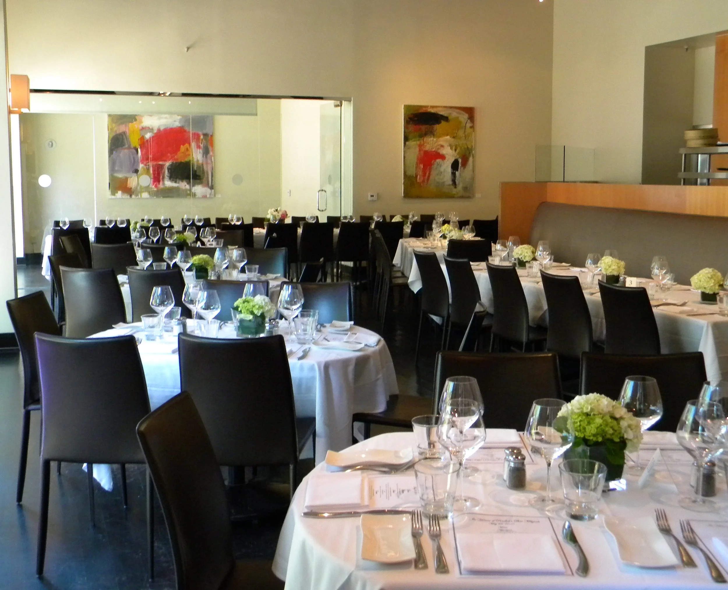A restaurant dining area set for a meal with white tablecloths, flower centerpieces, wine glasses, and silverware.