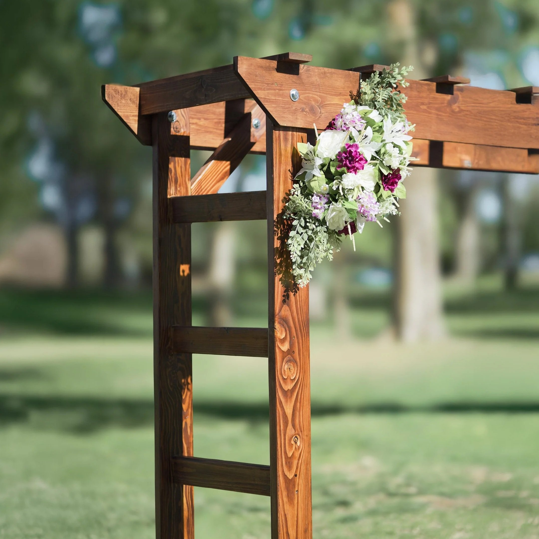 A wooden wedding arch decorated with white, pink, and purple flowers and green foliage set outdoors with a blurred background of trees and grass.