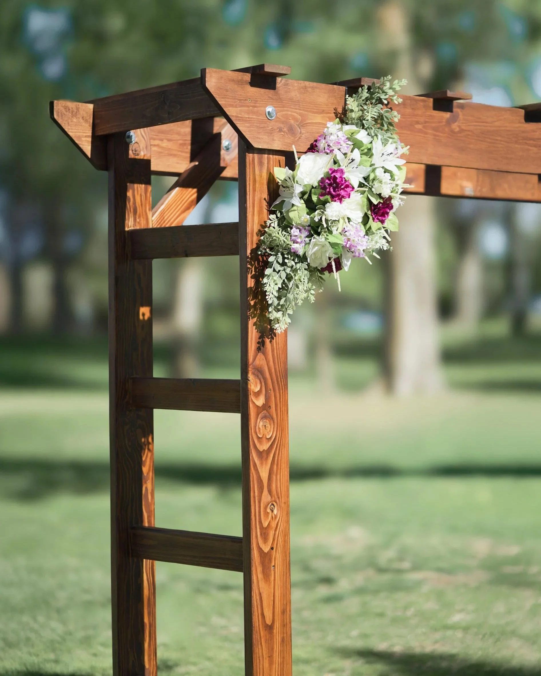 Wooden archway decorated with a floral arrangement of white, purple, and green flowers, set outdoors with trees and grass in the background.