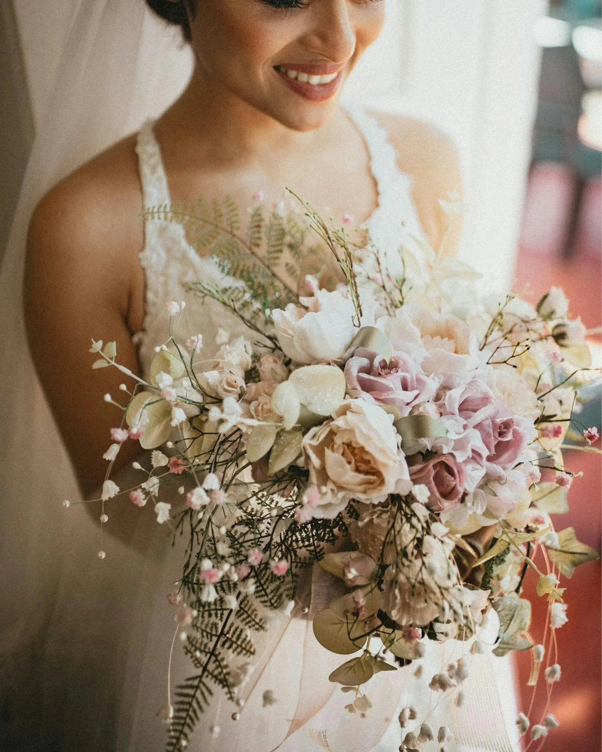 A smiling woman with short dark hair, wearing a white lace dress, holding a large bouquet of pink and white flowers with greenery.