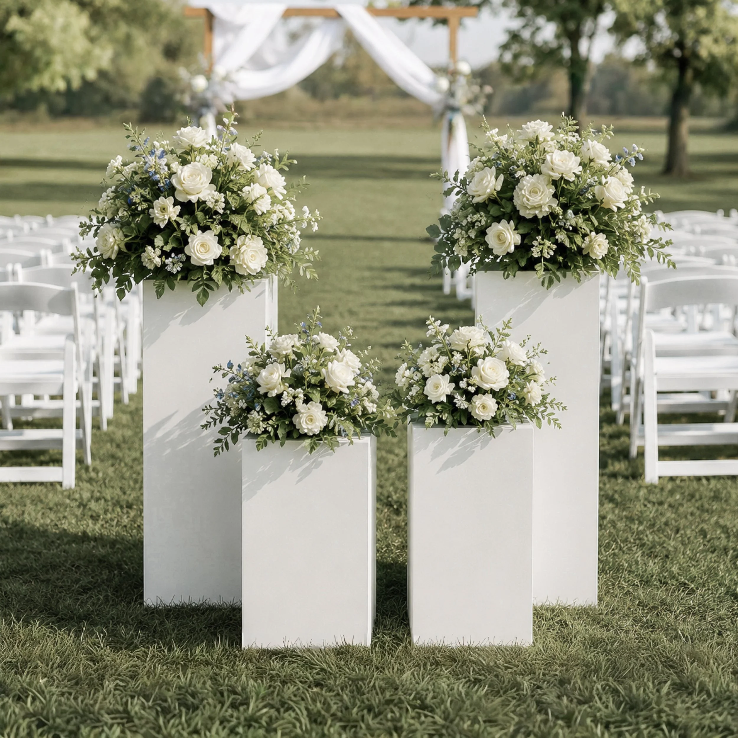 Two floral arrangements with white roses and green leaves on white podiums, outdoor wedding ceremony setup with chairs, an arch with drapery in the background, green trees, and grass.