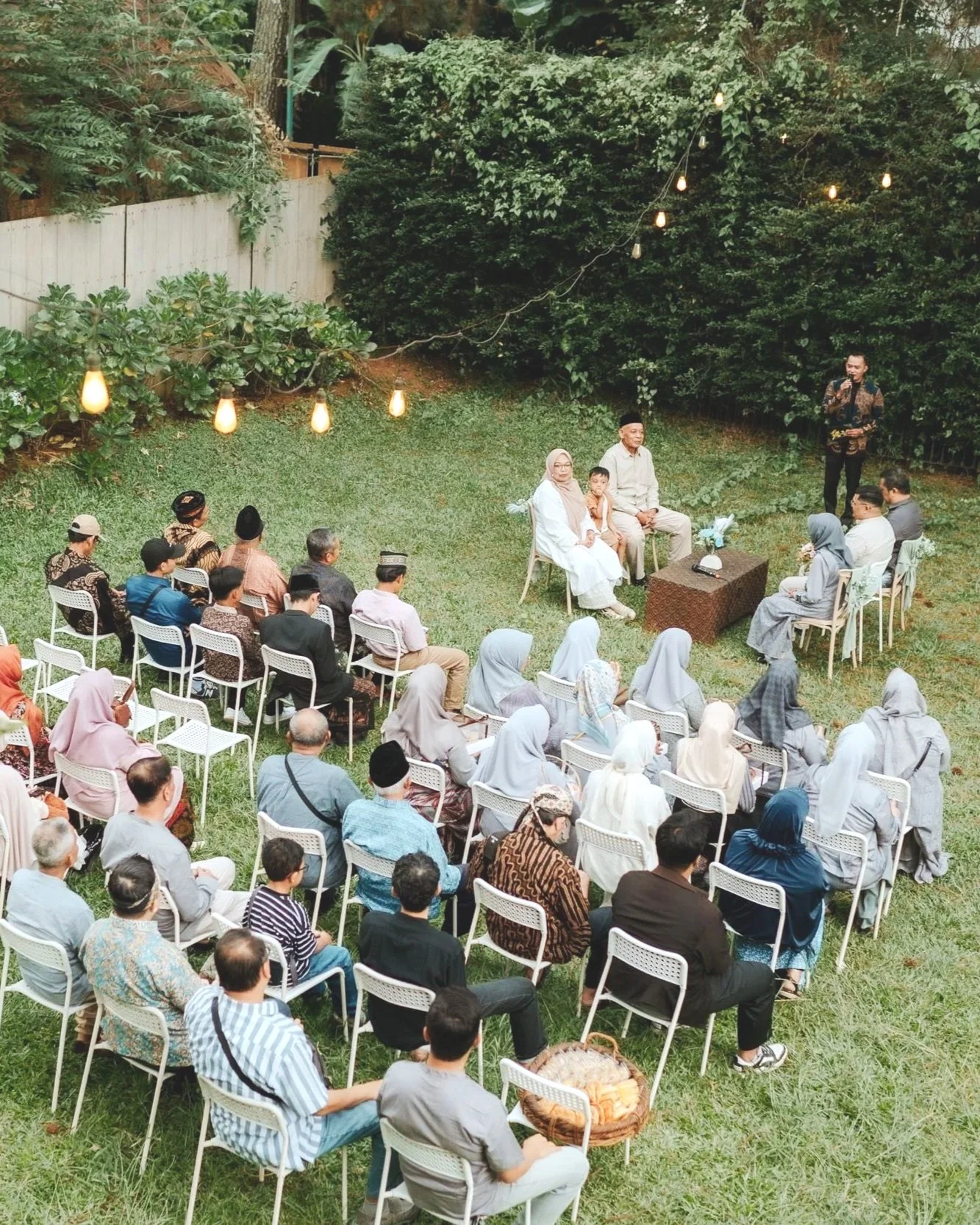 An outdoor gathering with multiple rows of chairs occupied by diverse attendees, a small stage with speakers, and a man speaking into a microphone, all set in a garden with string lights and greenery.