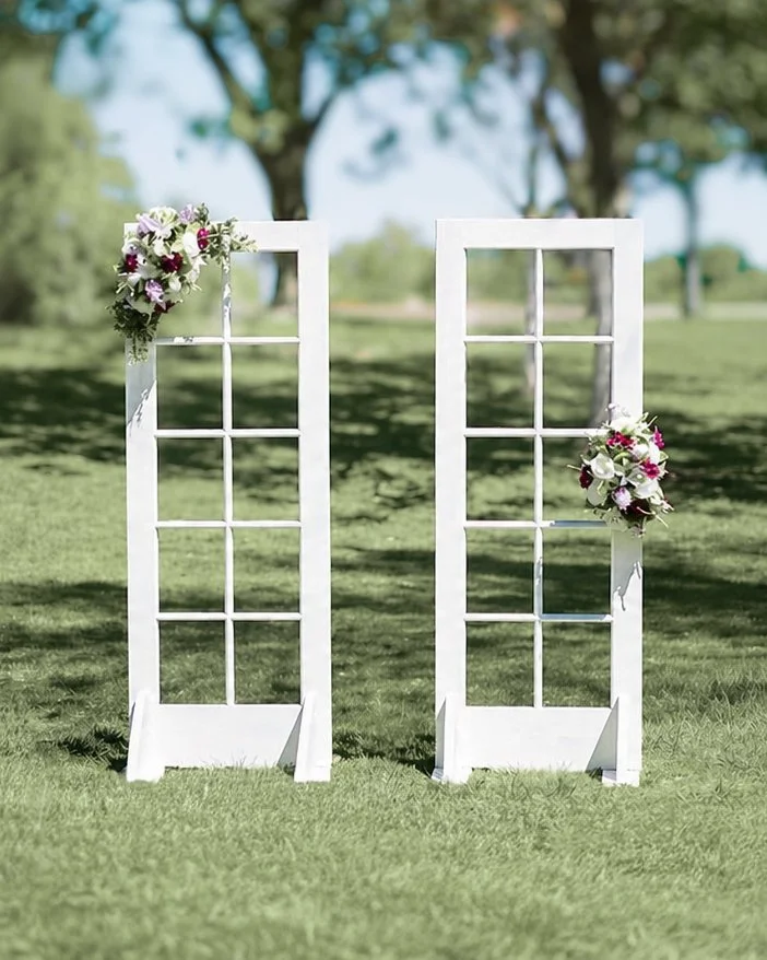 Two white decorative window frames with flower arrangements on a grassy outdoor setting.