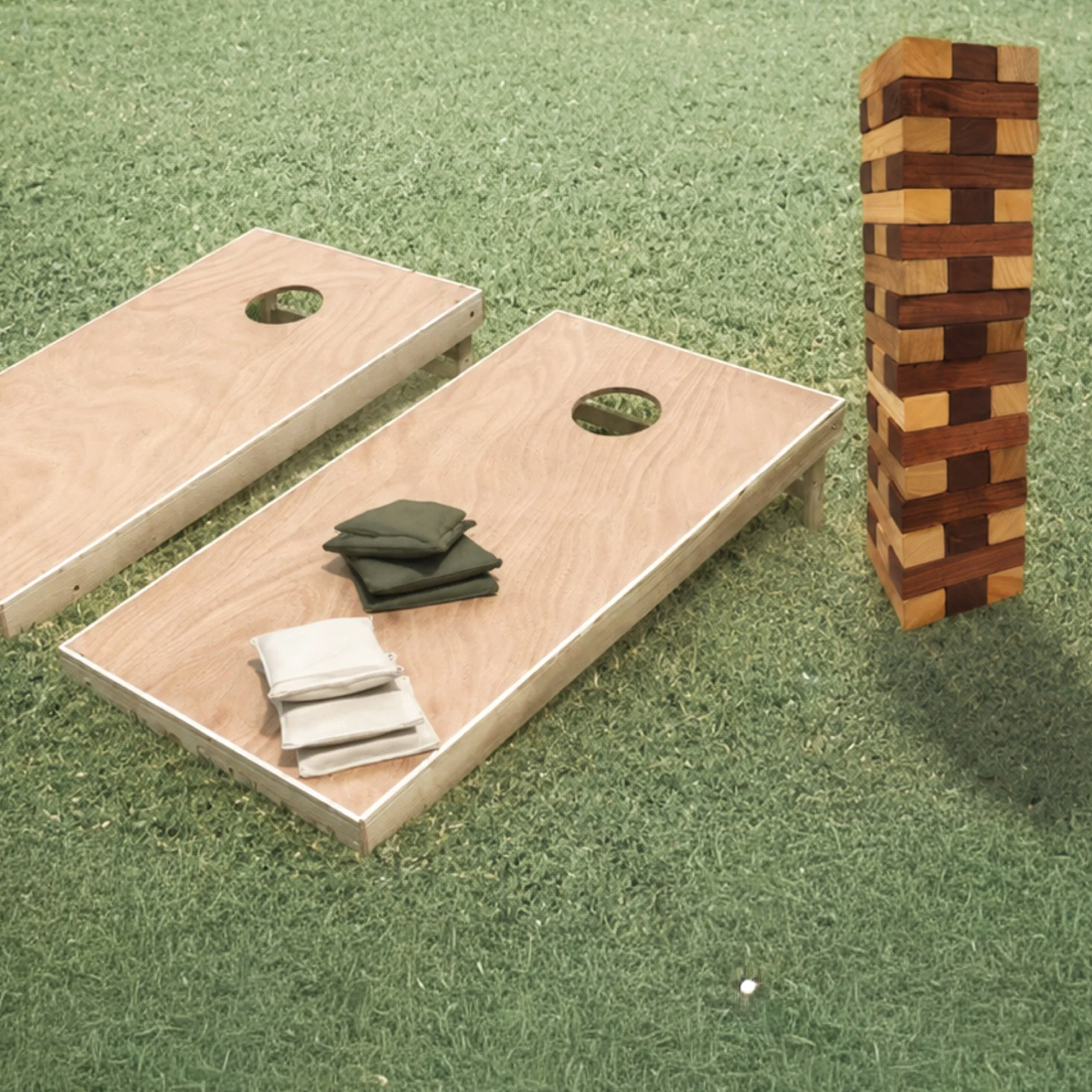 Two wooden cornhole game boards with holes on one side, a Jenga tower made of alternating light and dark wooden blocks, and a partially visible golf ball on grass.