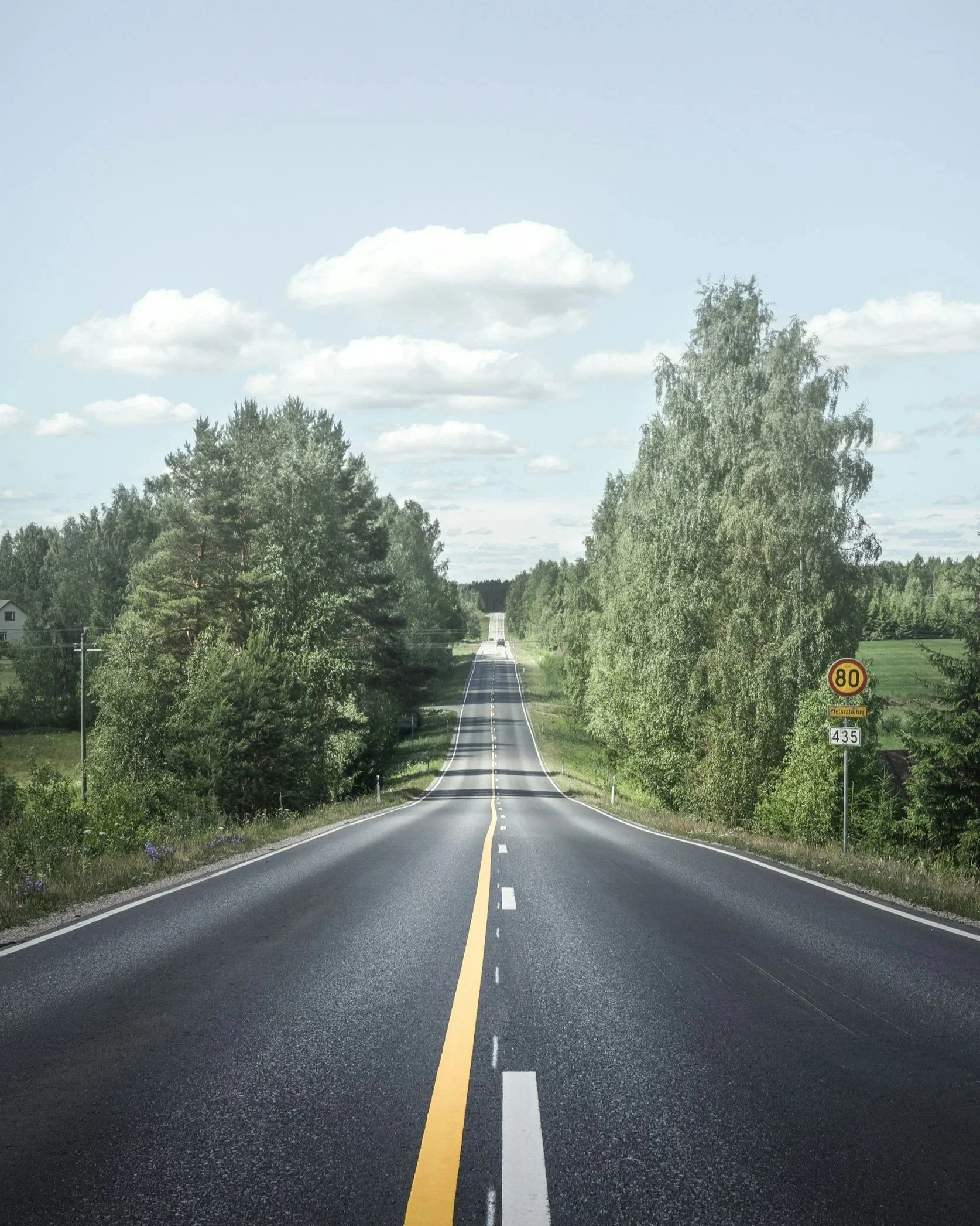 Open road stretching into the distance, flanked by green trees and a blue sky with a few clouds.