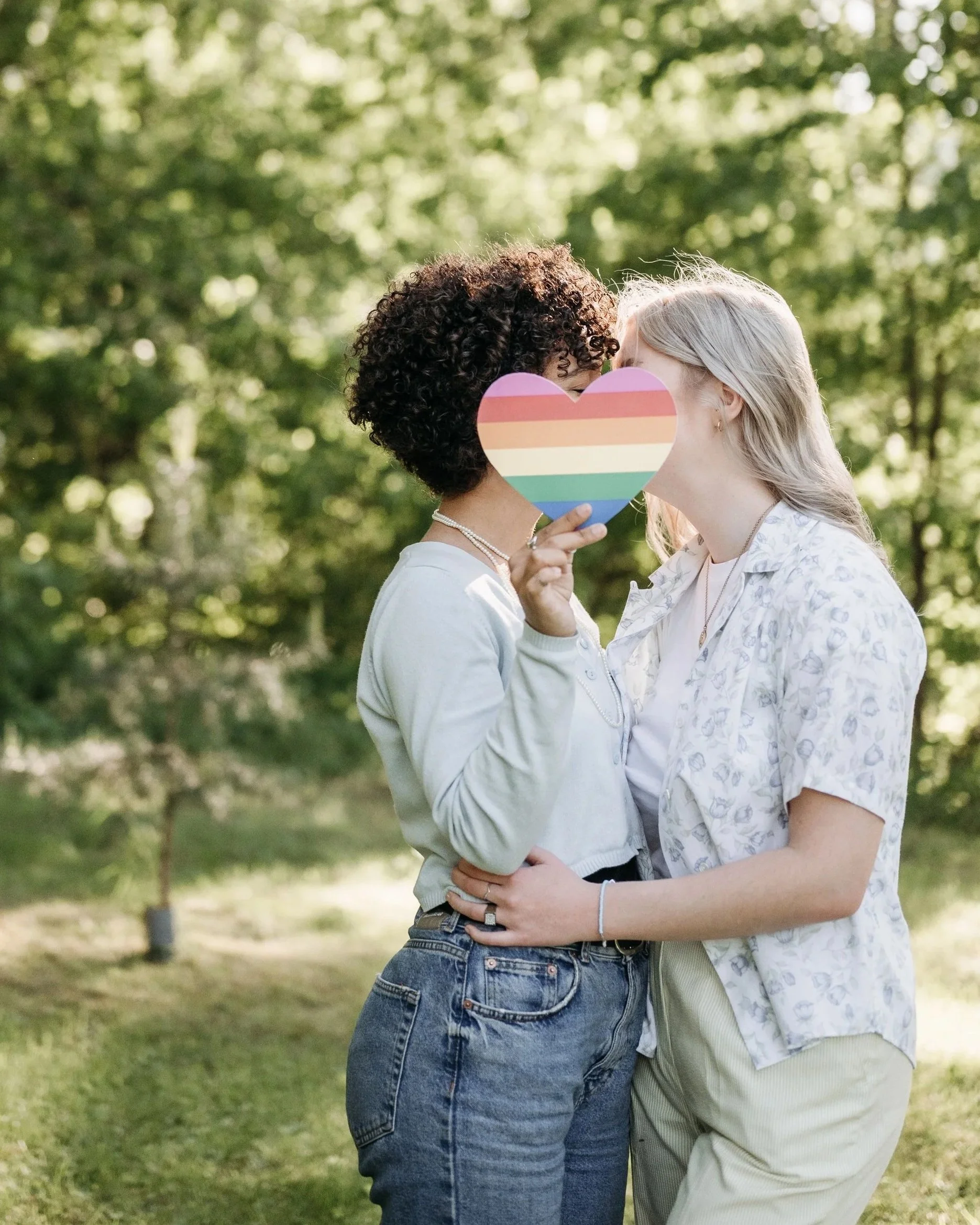 Two people are standing close together outdoors, holding a rainbow-colored heart-shaped paper in front of their faces. They are hugging and appear to be sharing an intimate moment. The background is a blurred green park with trees and sunlight.