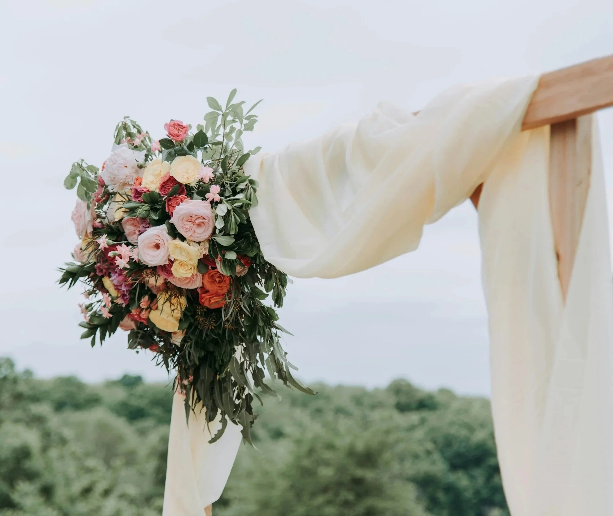 A bouquet of pink, yellow, orange, and white roses and assorted greenery attached to a wooden wedding arch draped with white fabric, set outdoors with a blurred green landscape background.