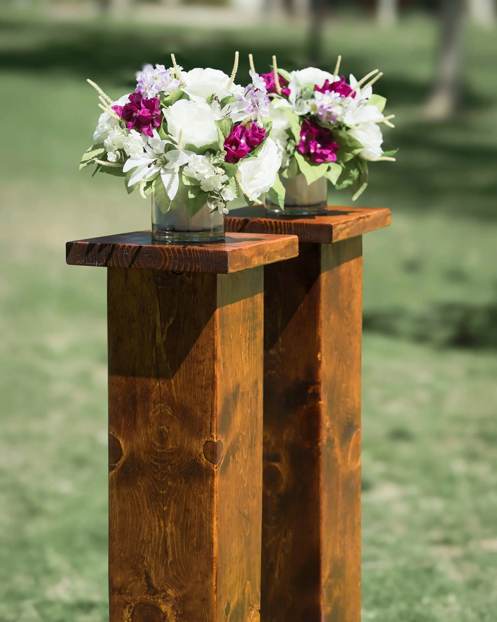 Two flower arrangements in glass vases on a wooden platform outdoors, with purple, white, and magenta flowers.