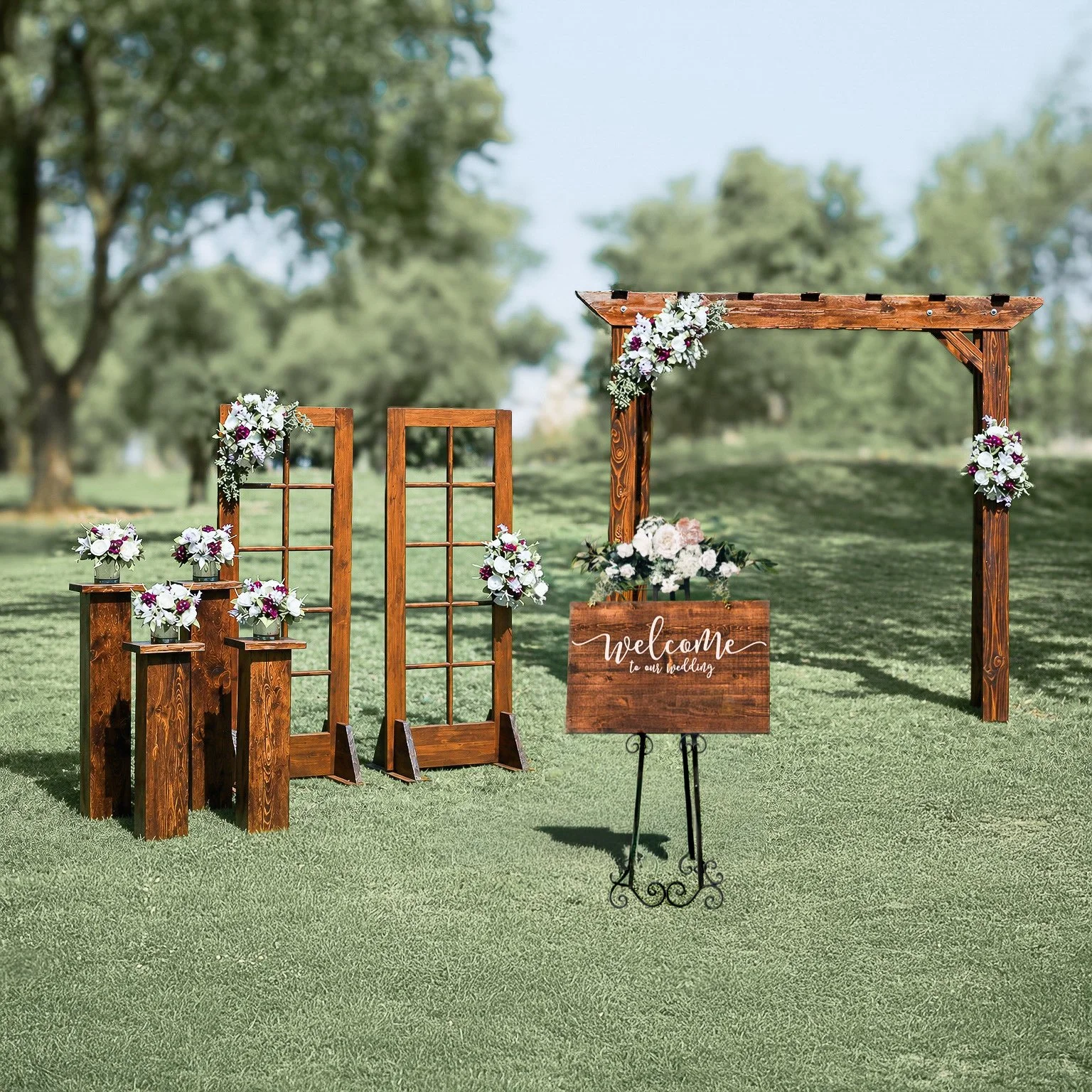 Outdoor wedding altar with a wooden arch decorated with flowers, a welcome sign, and small floral arrangements on wooden stands on a grassy lawn with trees in the background.