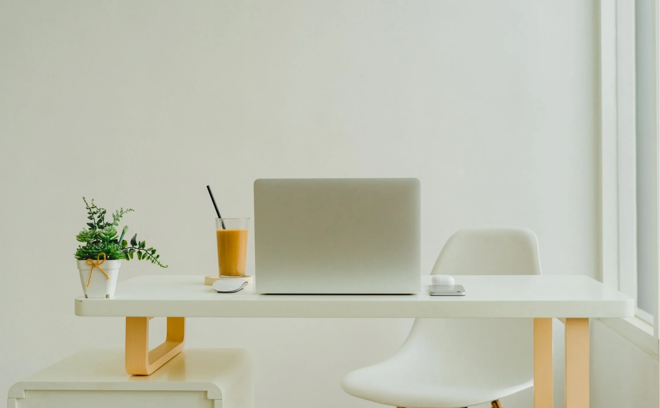 Minimalist home workspace with white desk, laptop, plant, glass of iced coffee, and white chair.
